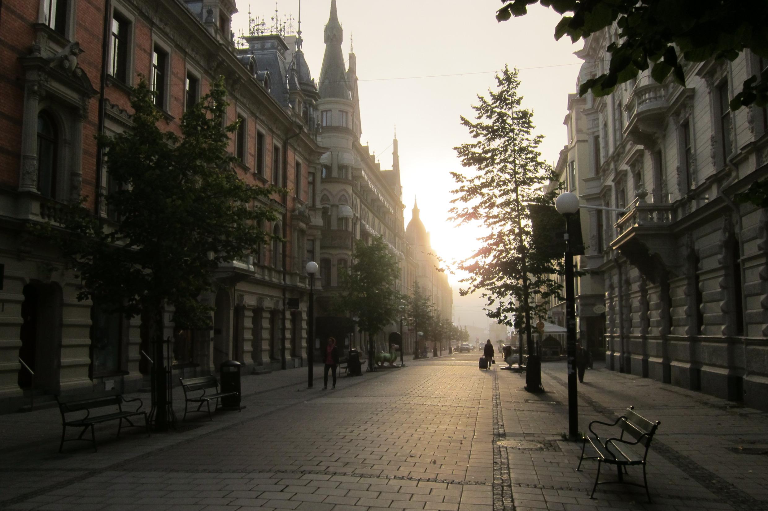 The sun shines at the end of a street in the stone city. A lone person with a suitcase is walking on the street.