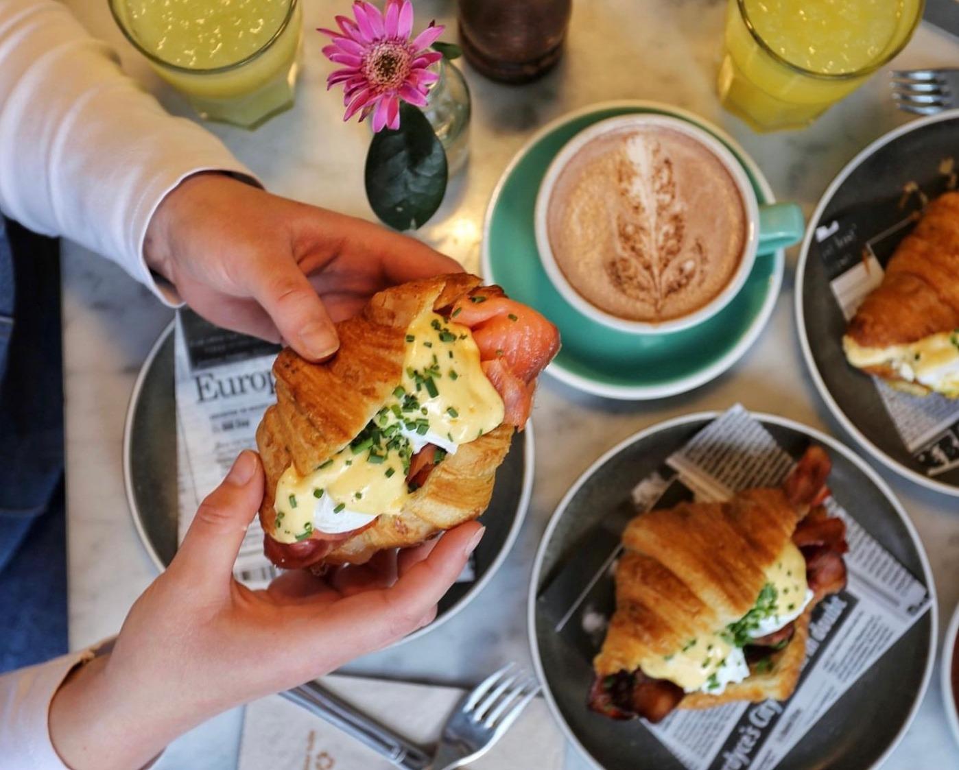 Une personne tient un croissant avec des œufs Bénédicte. Sont posées sur la table dessous, des assiettes avec des croissants, des verres de jus de fruits et un cappucino.