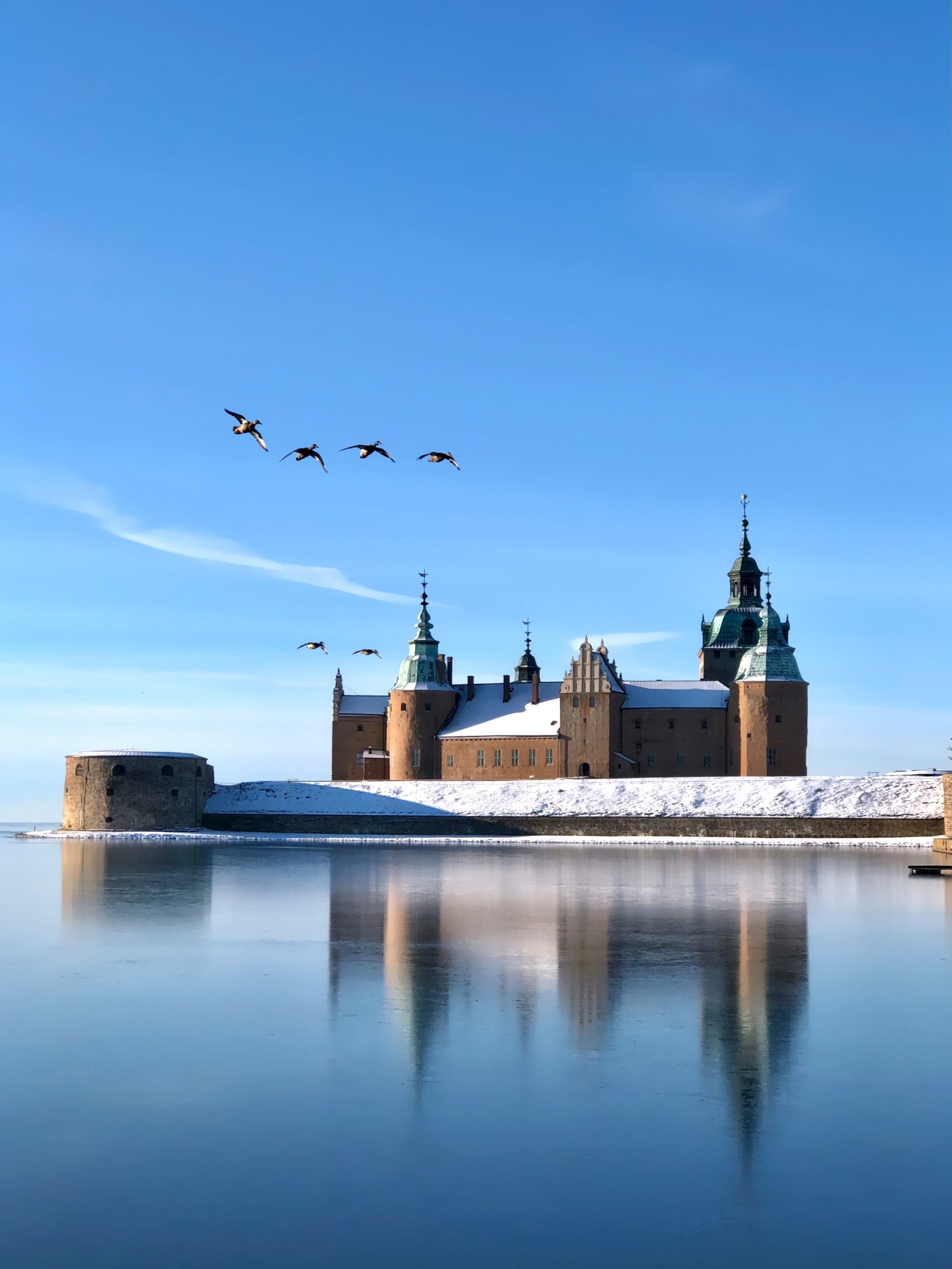 Le château de Kalmar recouvert de neige se reflète dans les douves gelées sous un ciel bleu clair d'hiver, avec des oiseaux volant au-dessus.