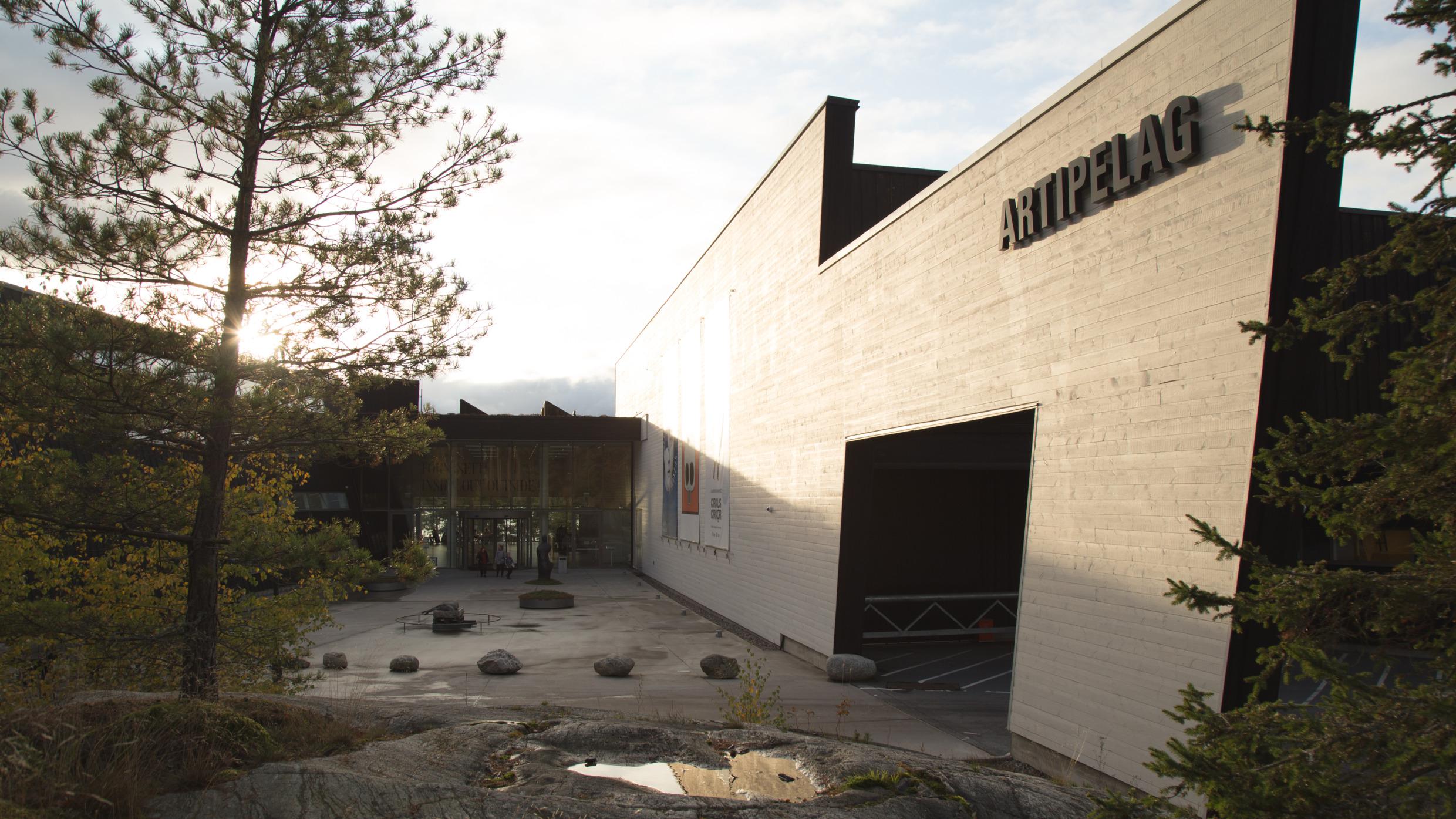 The house and the entrance to the Artipelag. On a large wood wall to the left you see a big sign that says ARTIPELAG. In front of the entrance is a yard with sculptures and some people come through the door. There is a tree in the front to the left.