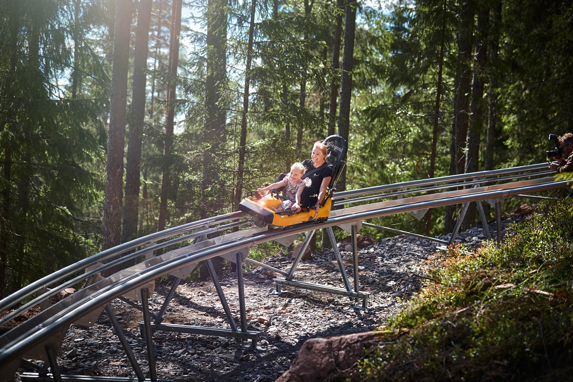 Een volwassene en een kind rijden in een alpine coaster door het bos bij Isaberg Mountain Resort, beiden glimlachen terwijl ze over de baan scheuren.