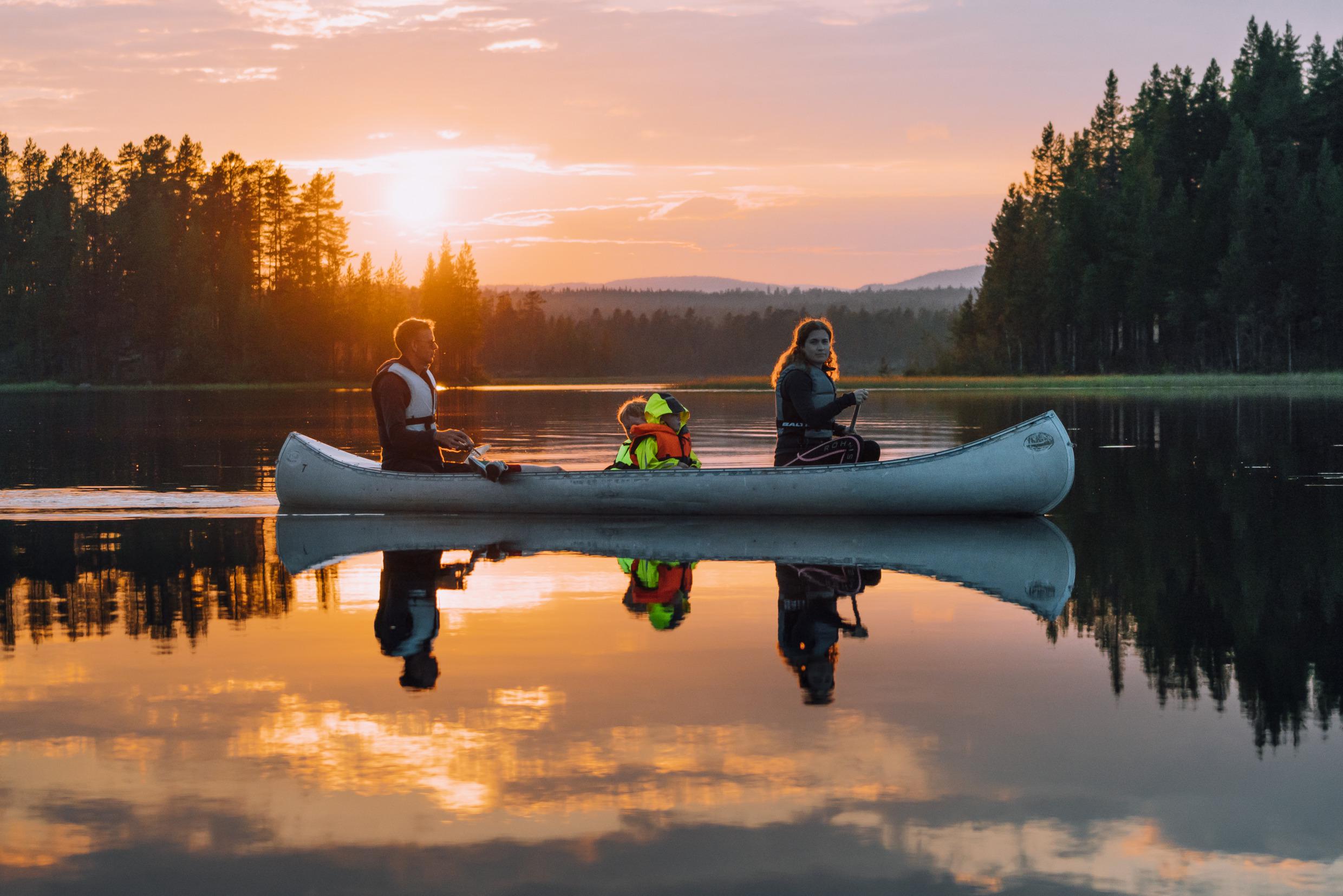 A family are canoeing in a lake surrounded by forest. The sun is setting in the horizon.