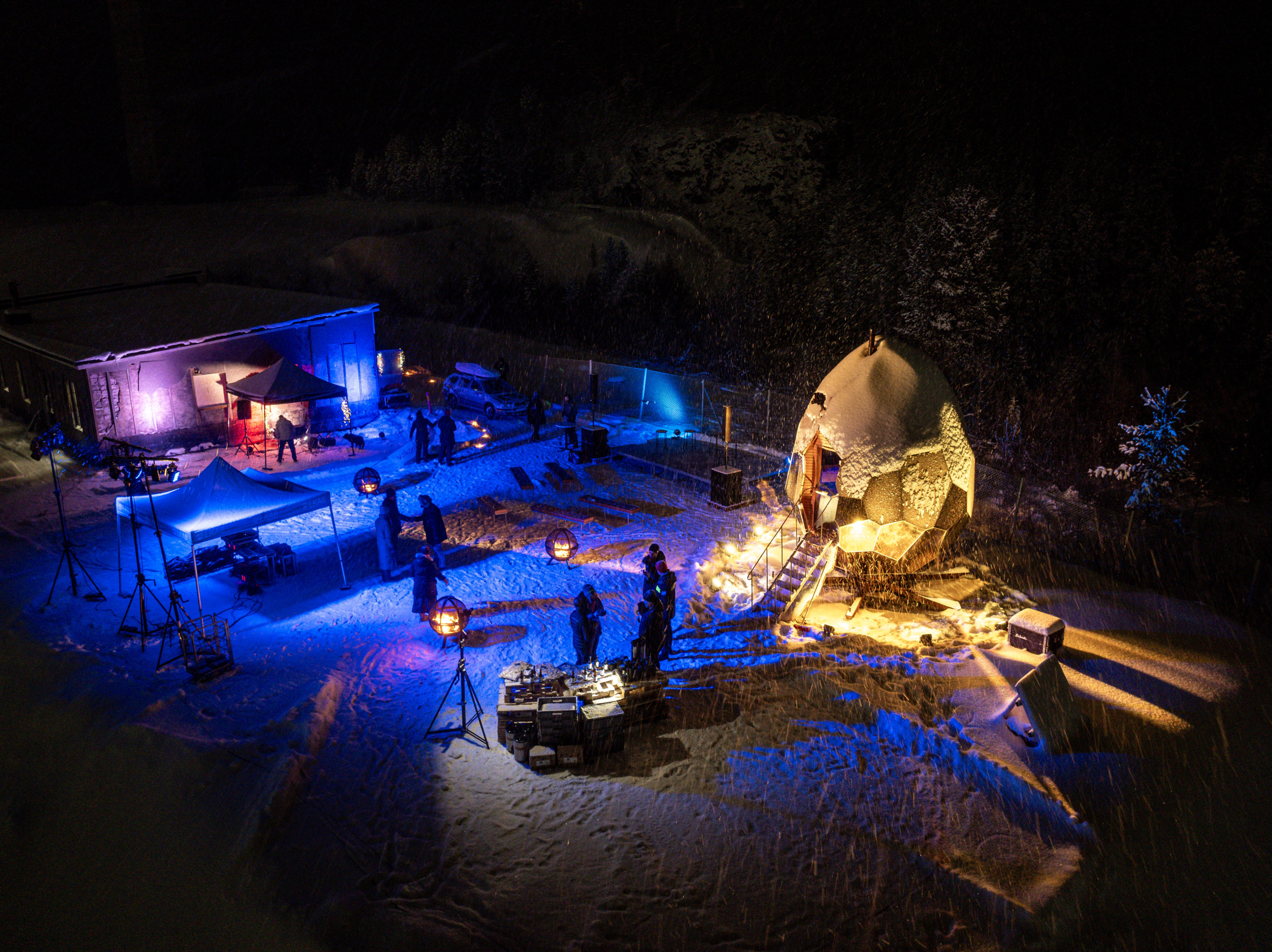 Nachtopname van een buitenevenement in het besneeuwde Kiruna, Zweden, met de verlichte gouden Solar Egg-sauna omringd door mensen en kleurrijke lampjes.