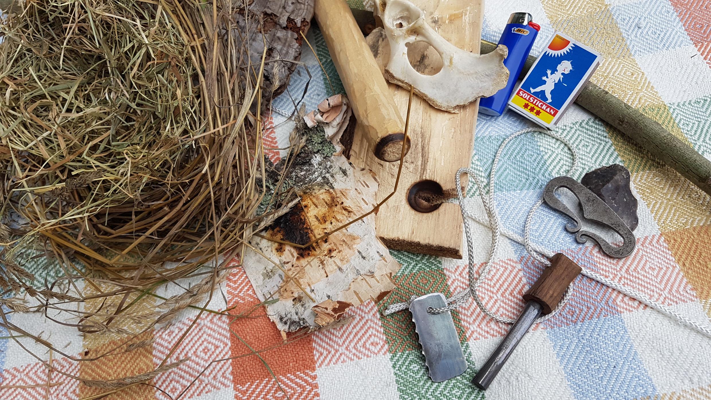 Auf einer Decke liegen Gegenstände, um ein Feuer zu machen. Es gibt ein Feuerzeug, Brennholz, Streichhölzer und Birkenrinde.