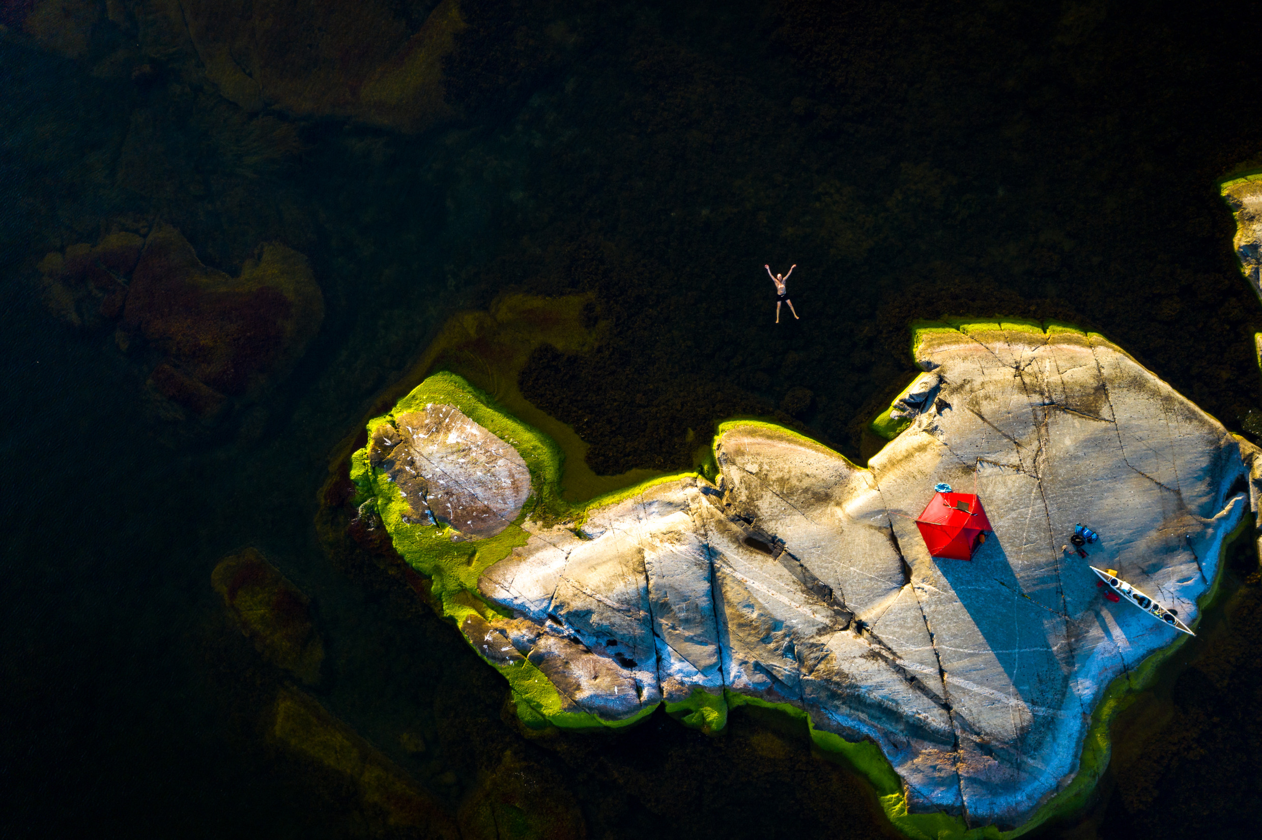 Drone footage of a man swimming in the archipelago. A red tent and a white kayak lie on the island next to the man.