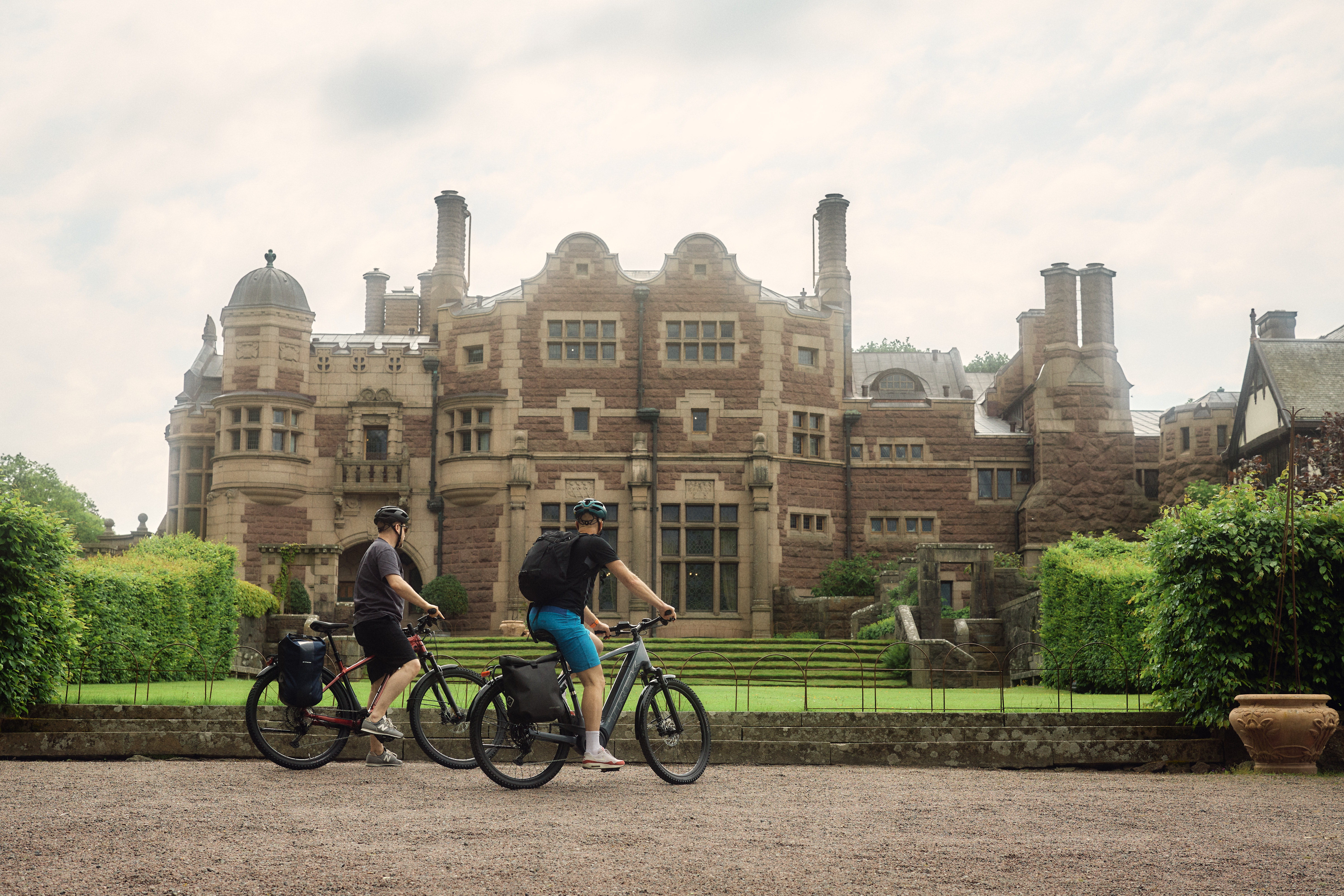 Two cyclists in front of Tjolöholm Castle, a Tudor-style stone castle surrounded by gardens and lush greenery on Sweden's west coast.