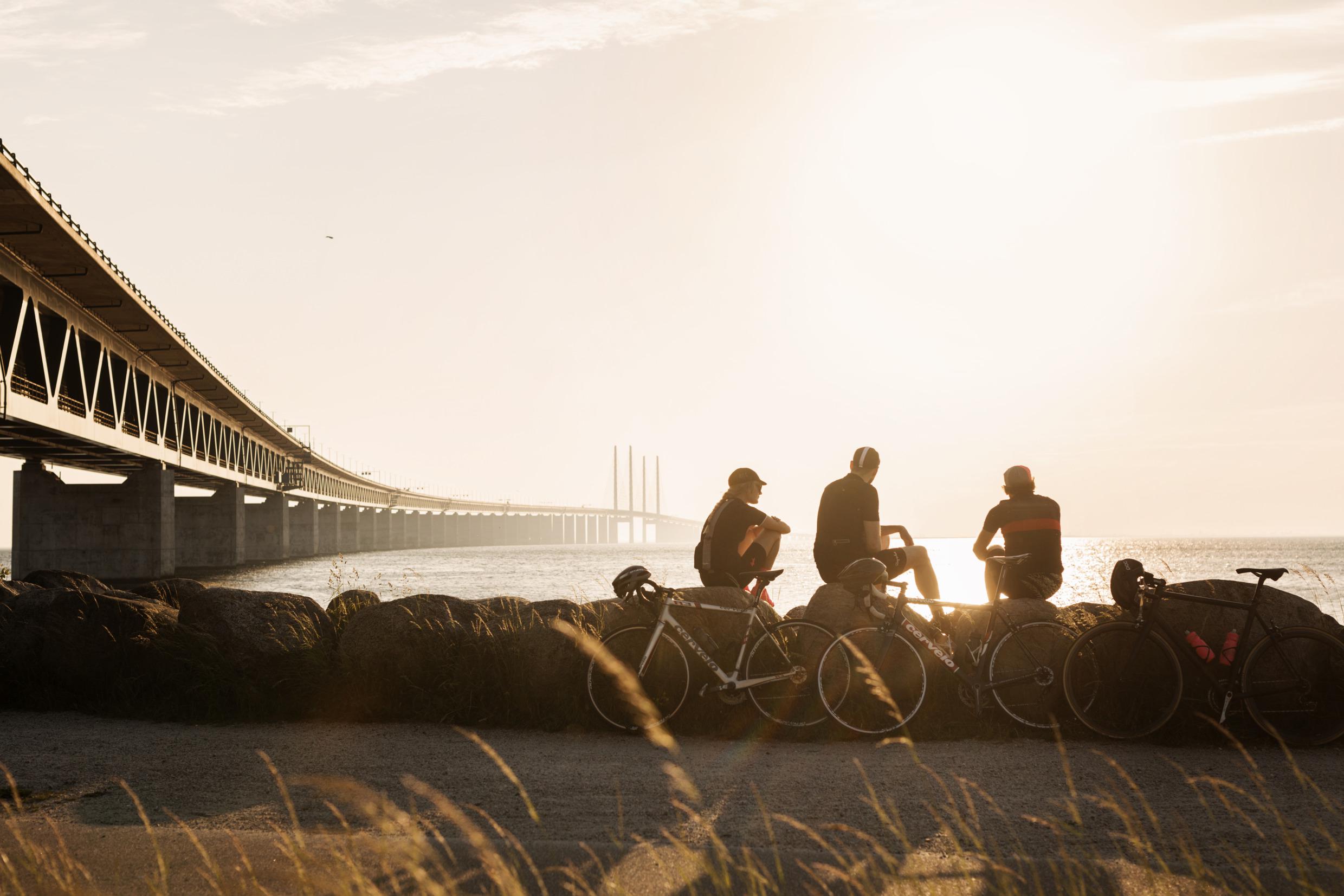 Blick auf die Brückenpilonen der Öresundbrücke. Drei Fahrradfahrer stehen auf einem Radweg vor der Brücke und beobachten den Sonnenuntergang