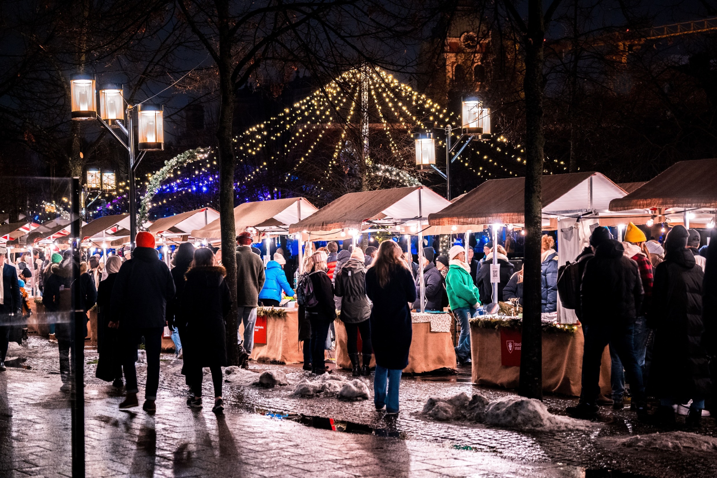 Mensen die rondkijken bij de vele kraampjes op de kerstmarkt in Kungsträdgården, Stockholm. Het is donker, maar de lichtjes en straatlantaarns maken het gezellig.