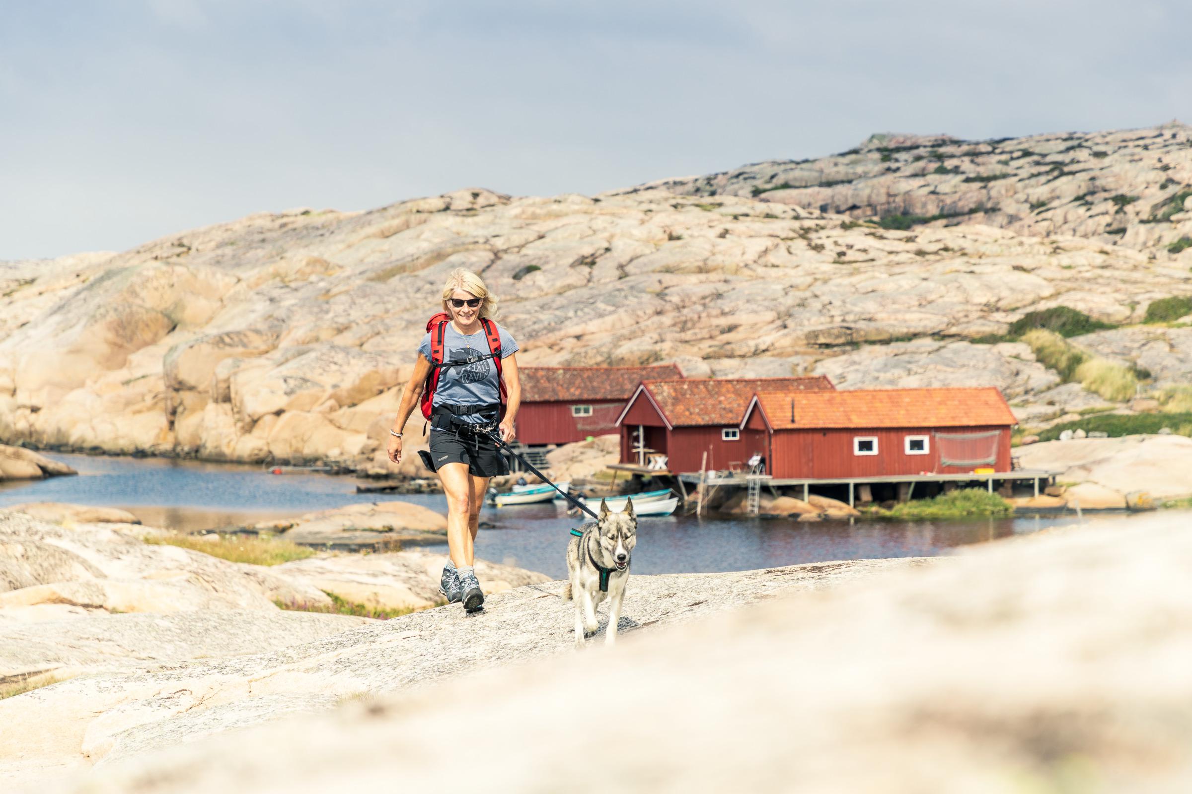 Eine Frau mit ihrem Hund an der Leine wandert auf einem Pfad auf Raketen in der Nähe des Wassers. Bootshäuser am Wasser.