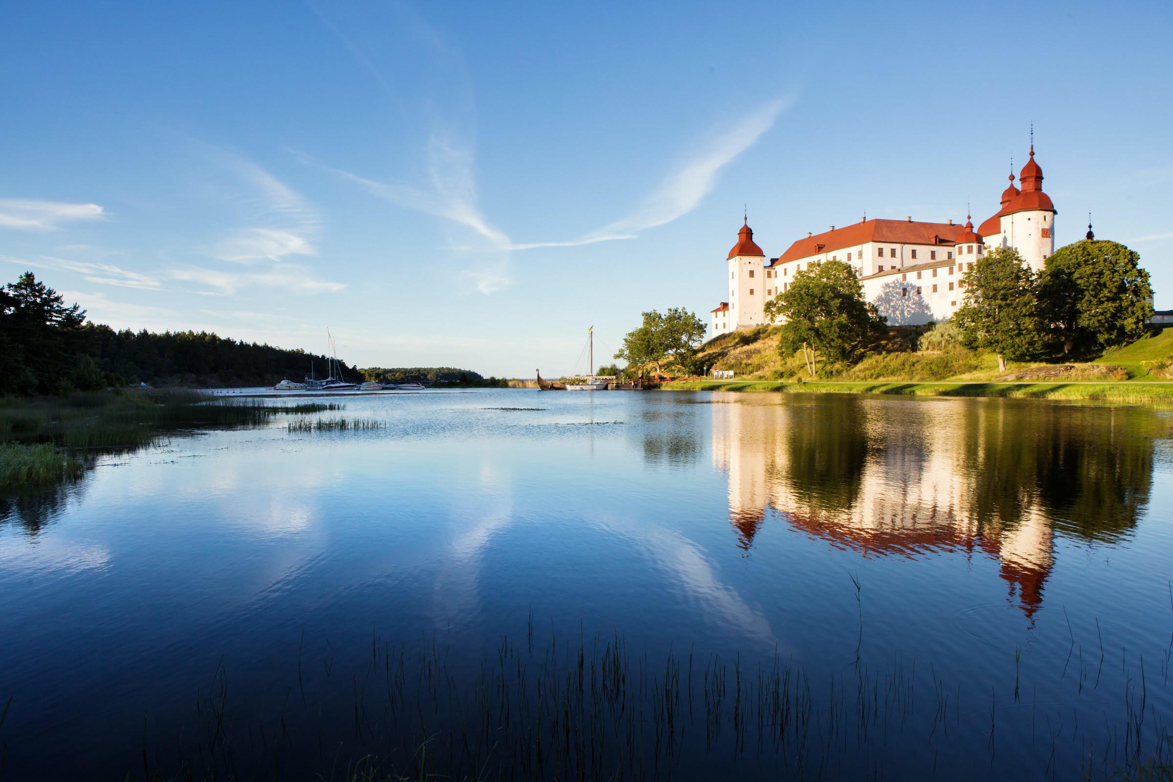 A large white castle with a red roof by a calm lake surrounded by greenery.