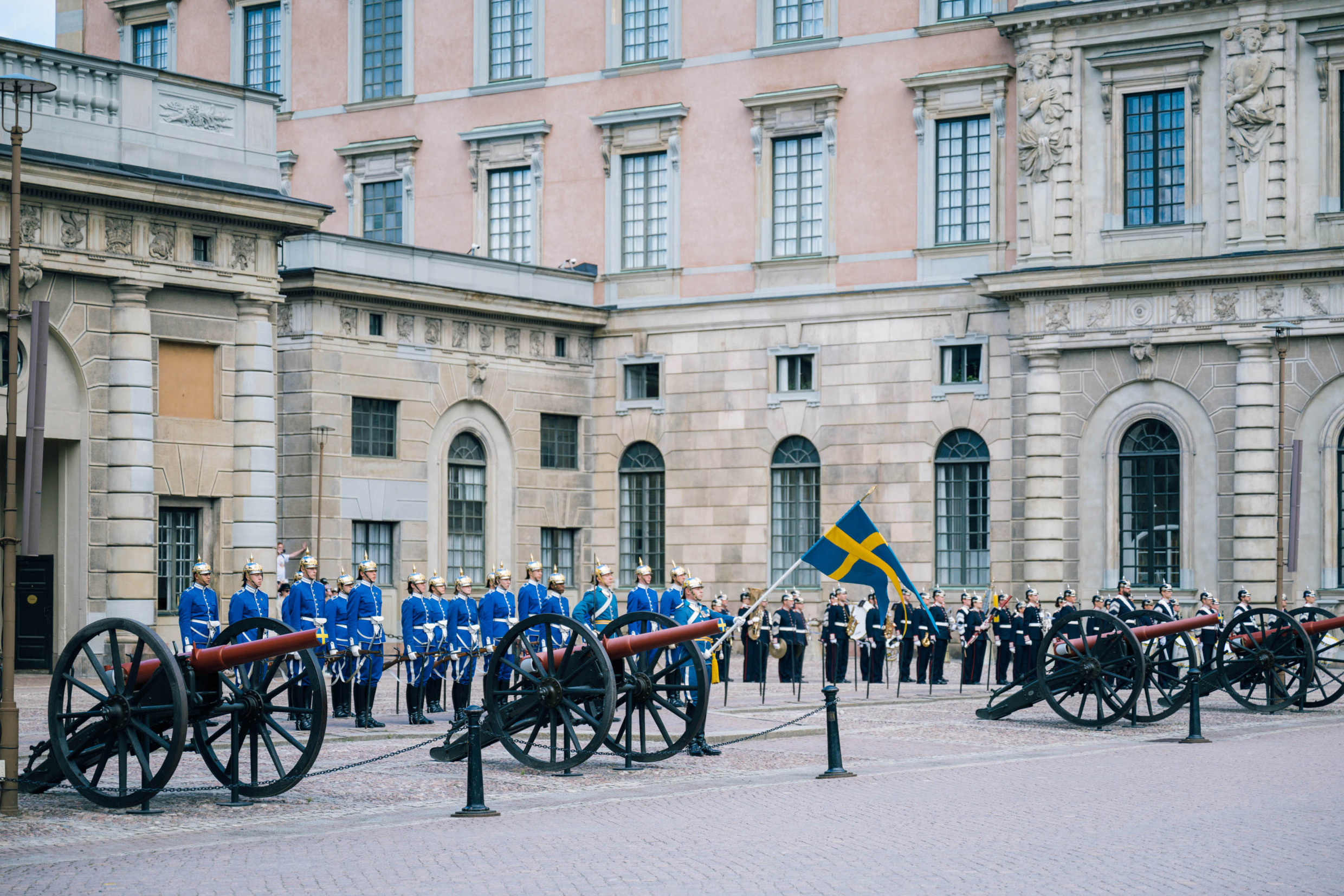 The royal guards outside the Royal Palace in Stockholm. They are holding a Swedish flag and next to them are antique cannons.