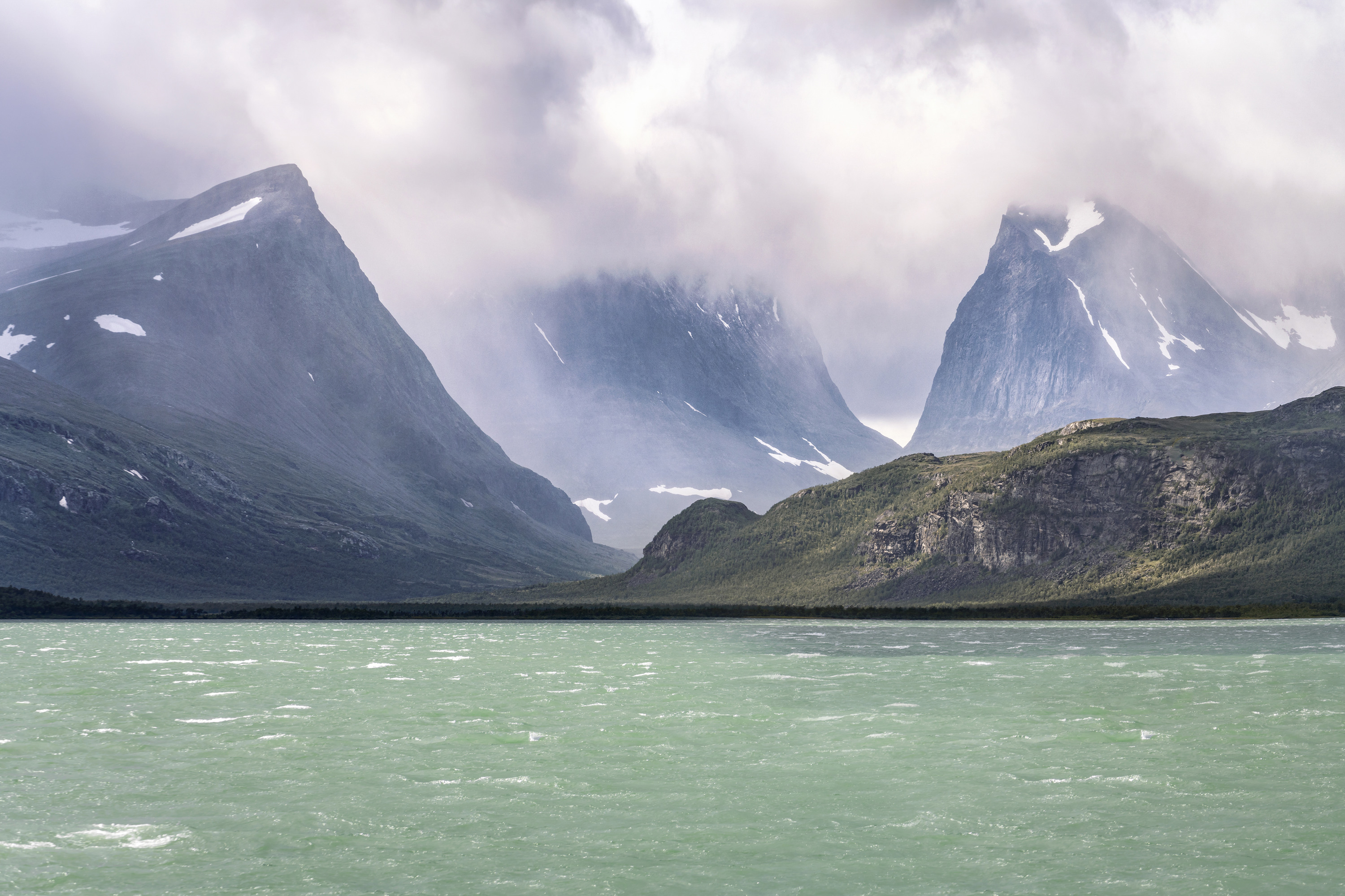 Ein türkisfarbener Bergsee liegt unter steilen, schroffen Gipfeln mit Schneeflecken, während tiefe Wolken zwischen den Bergen in Laponia dahinziehen.