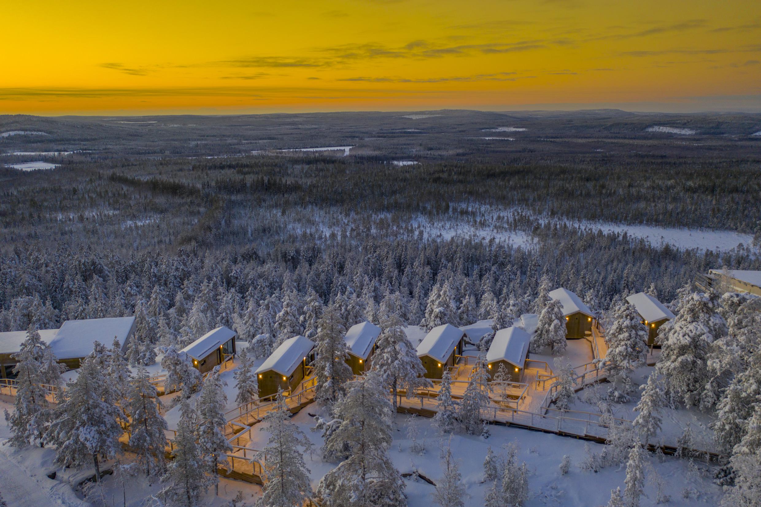 Vue aérienne d'une forêt enneigée et des chalets du Lapland View Lodge au coucher du soleil.
