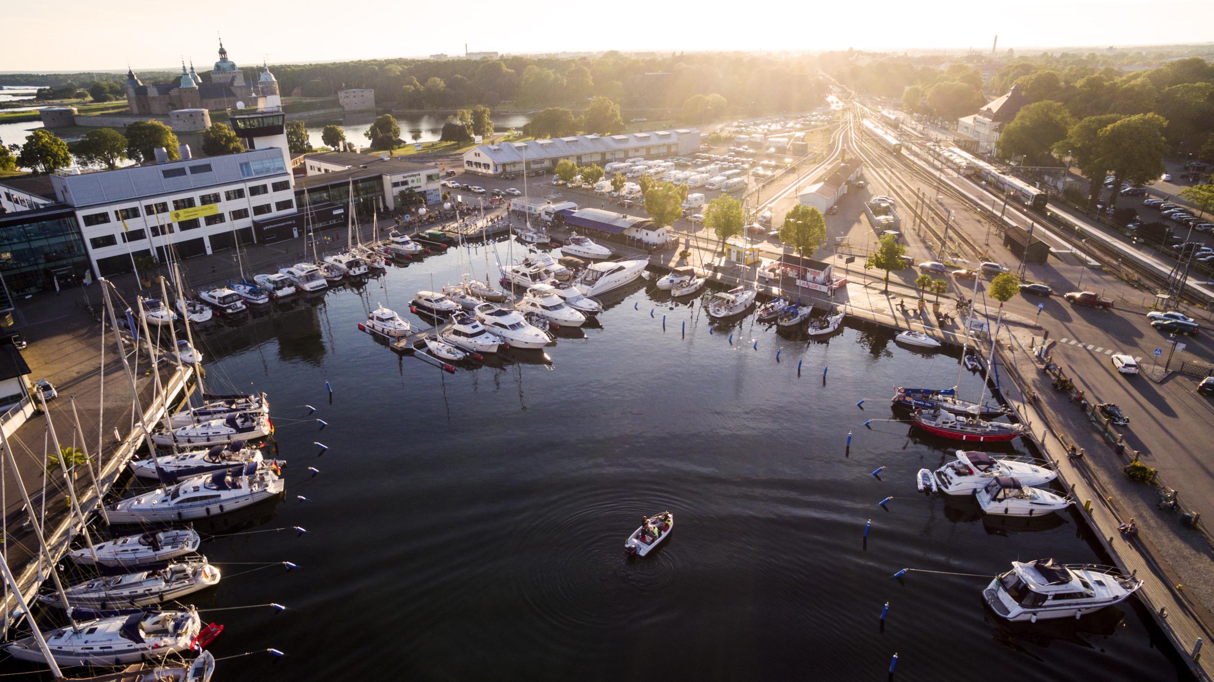 Luftaufnahme des Gästehafens von Kalmar mit mehreren Booten, die an den Stegen vor Anker liegen. Rechts ein Bahnhof und links das Schloss Kalmar im Hintergrund.