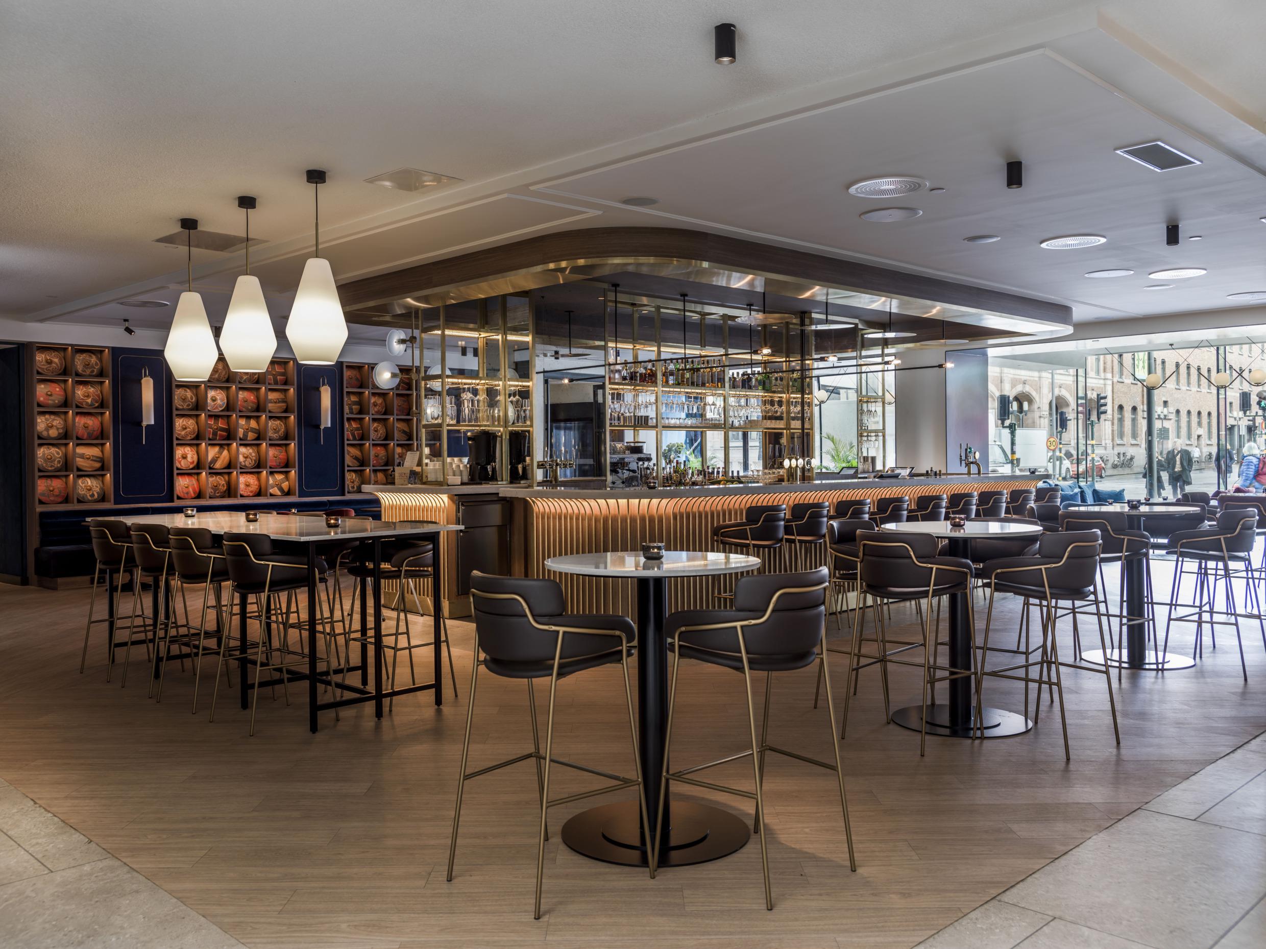 Een hotellobby met een bar in het midden. Rondom de bar staan bartafels en krukken.