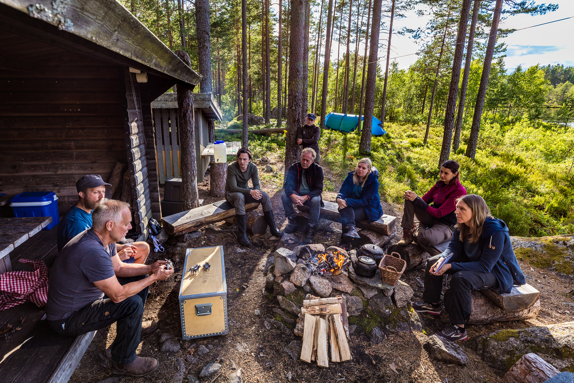 A group of people sitting around a campfire in a forest, listening to a guide and cooking outdoors.