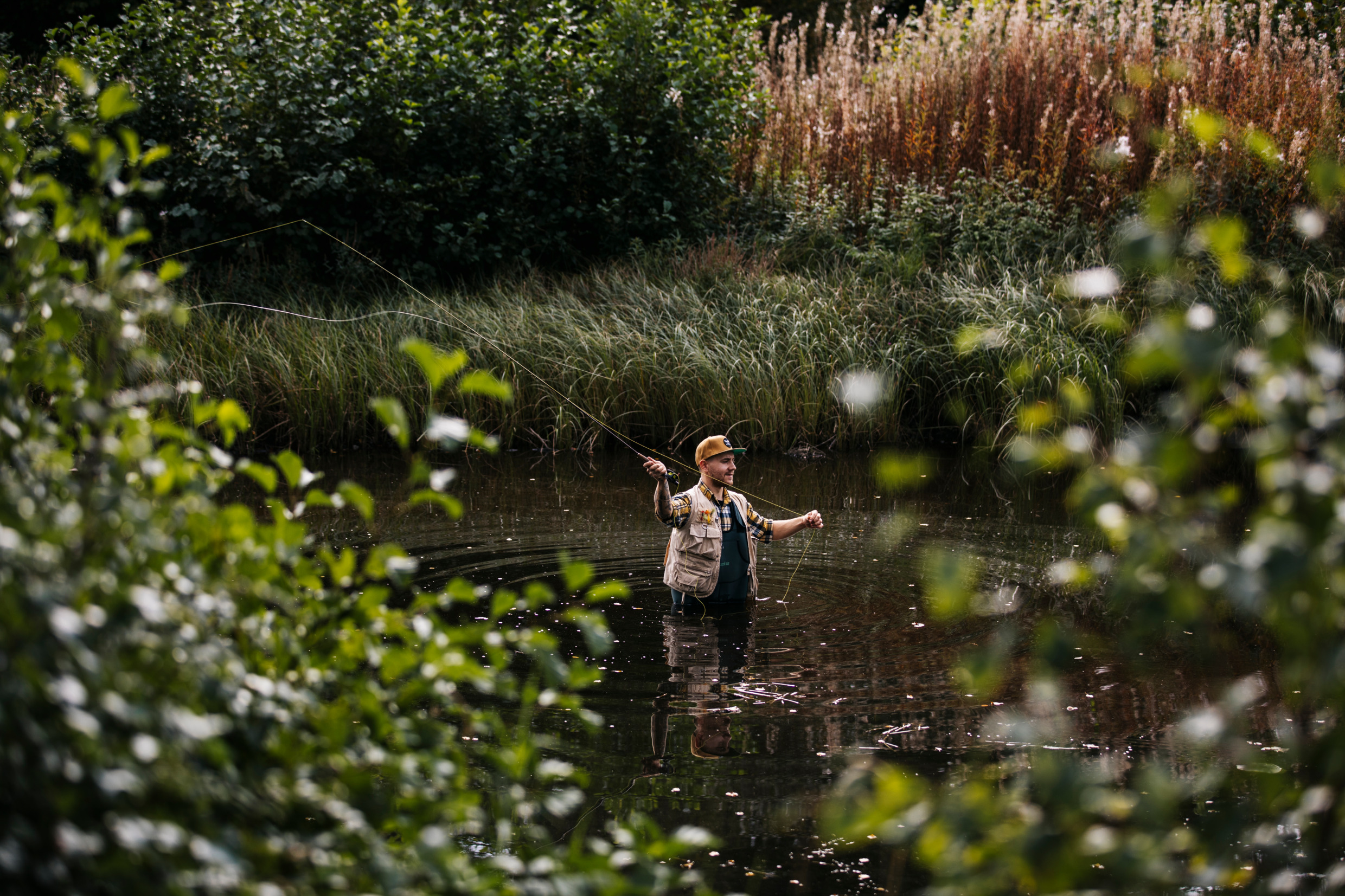 A man standing in a lake with water up to his hips, casting a fishing rod.