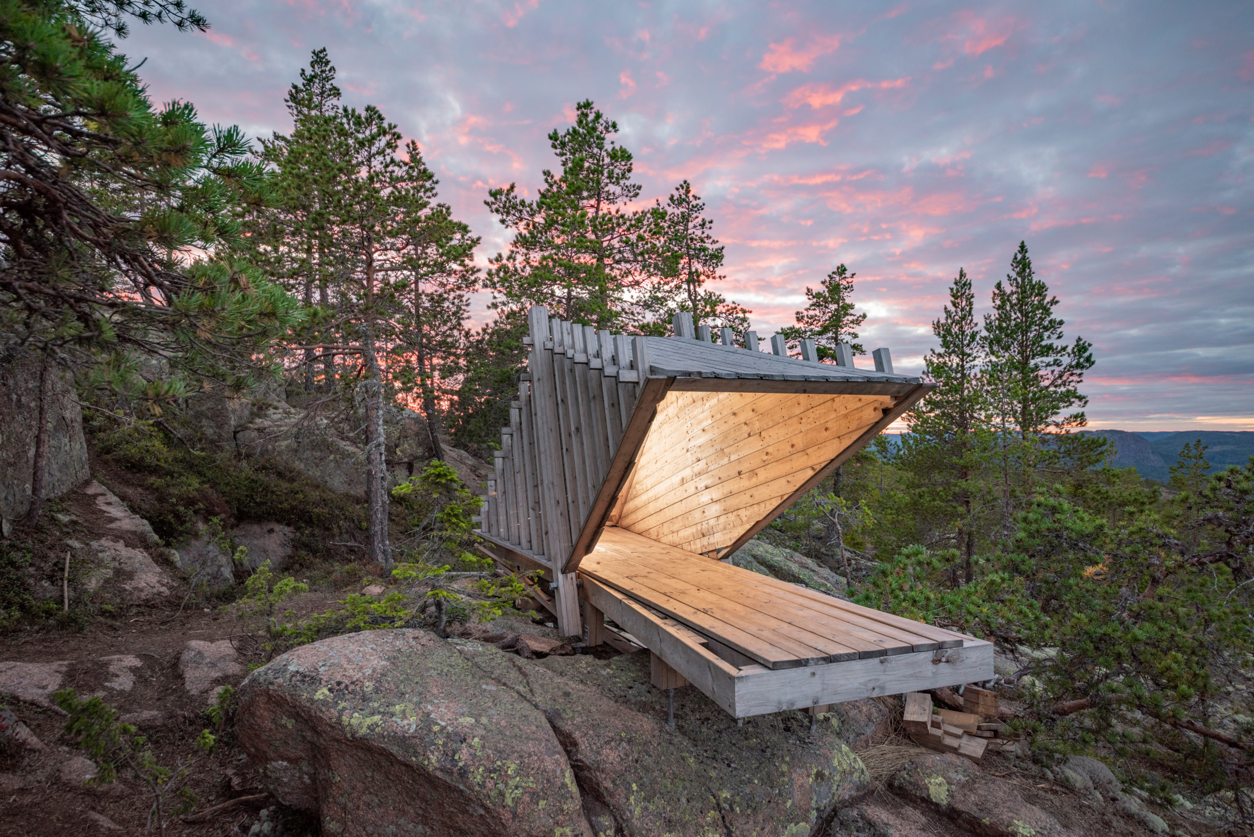 A hut or shack built with minimalist architecture stands on top of a forested hill of the High Coast of Sweden, with a twilight sky behind it.