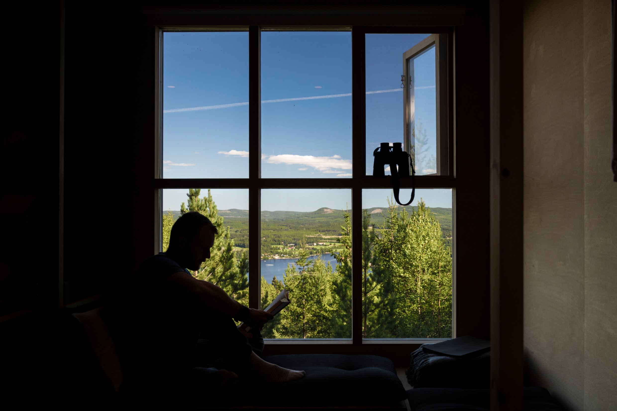 Silhouet van een man die een boek leest tegen een groot raam en uitkijkt over de blauwe lucht en de bergen erboven in Hälsingland, met een verrekijker in de aanslag.