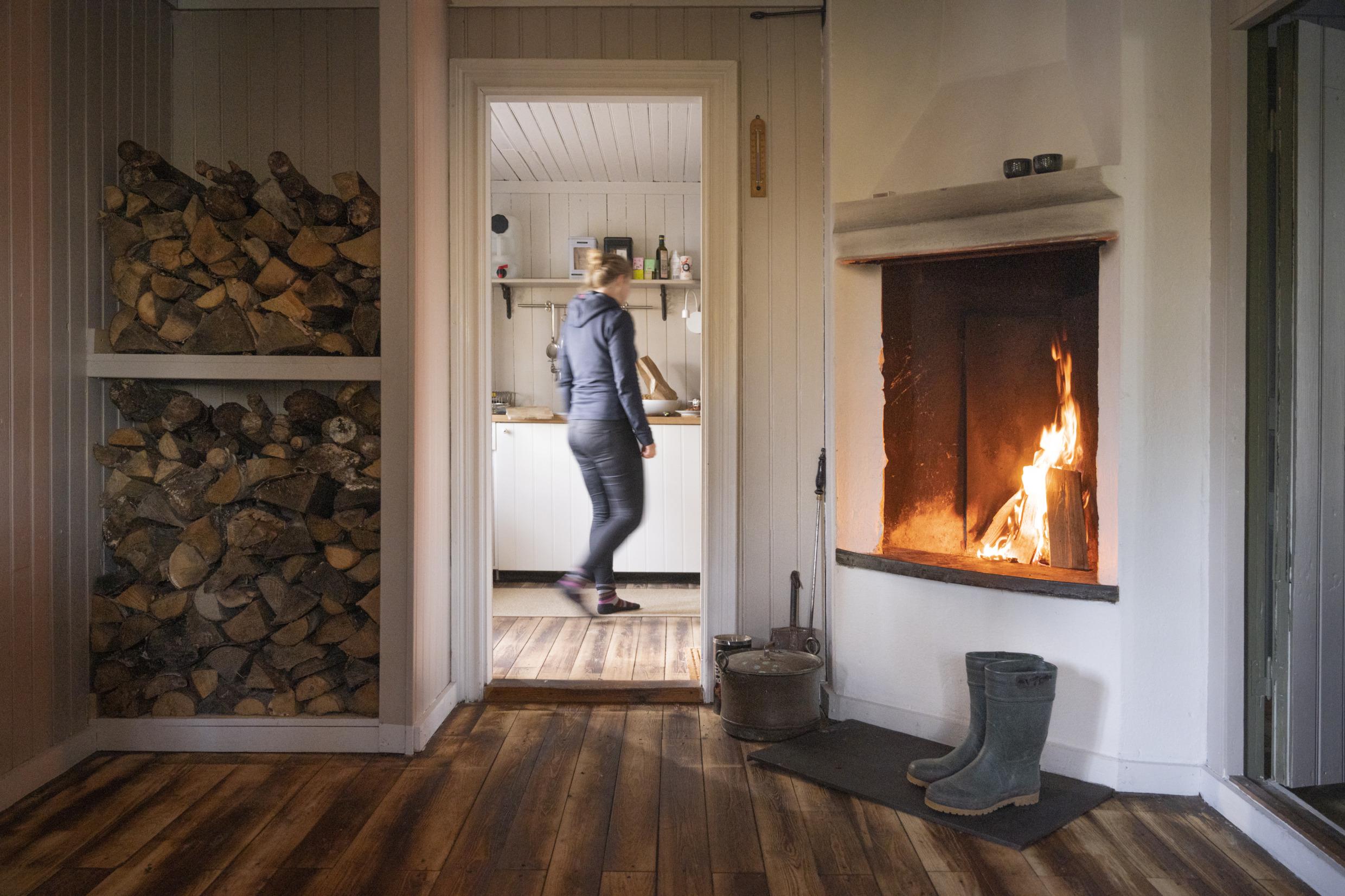 Ein Paar Gummistiefel stehen zum Trocknen unter einem knisternden Feuer im Kamin einer Hütte, während man die Silhouette einer Frau sieht, die durch den Raum nebenan geht.