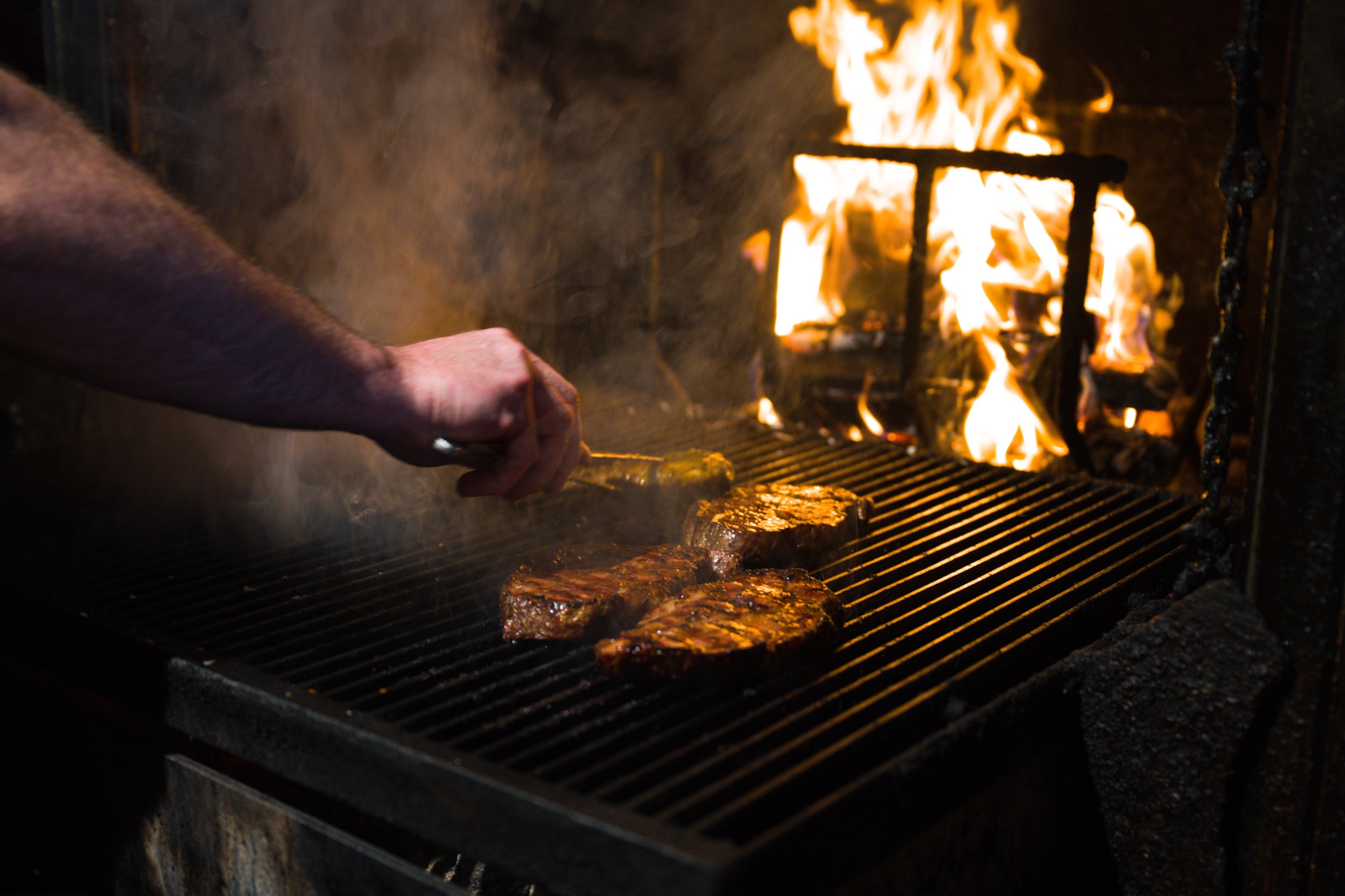Meat being handled on a grill with a fire in the background.