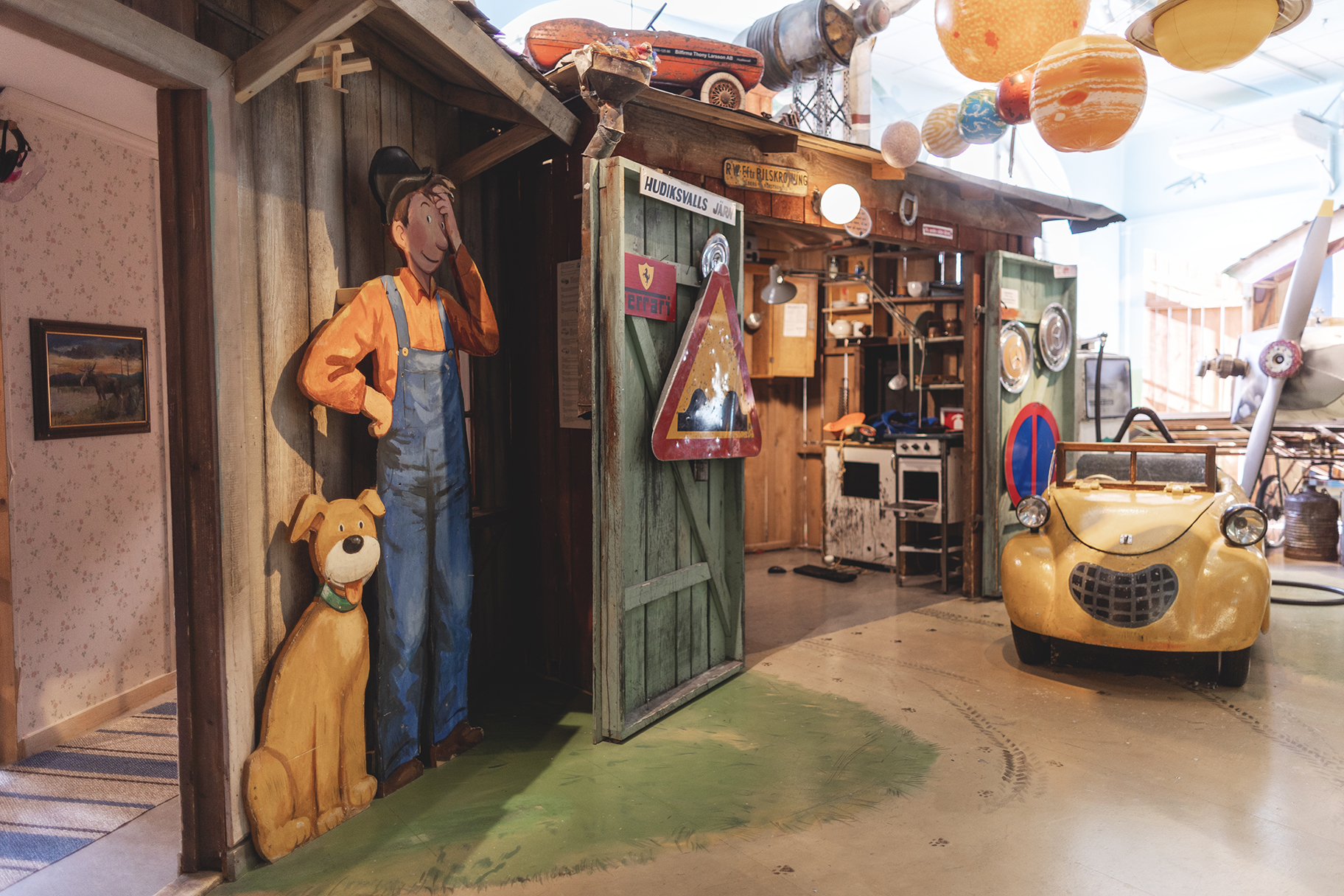 Indoor children’s exhibit with cartoon figure Mulle Meck, his dog and a yellow toy car, surrounded by playful details and tools.