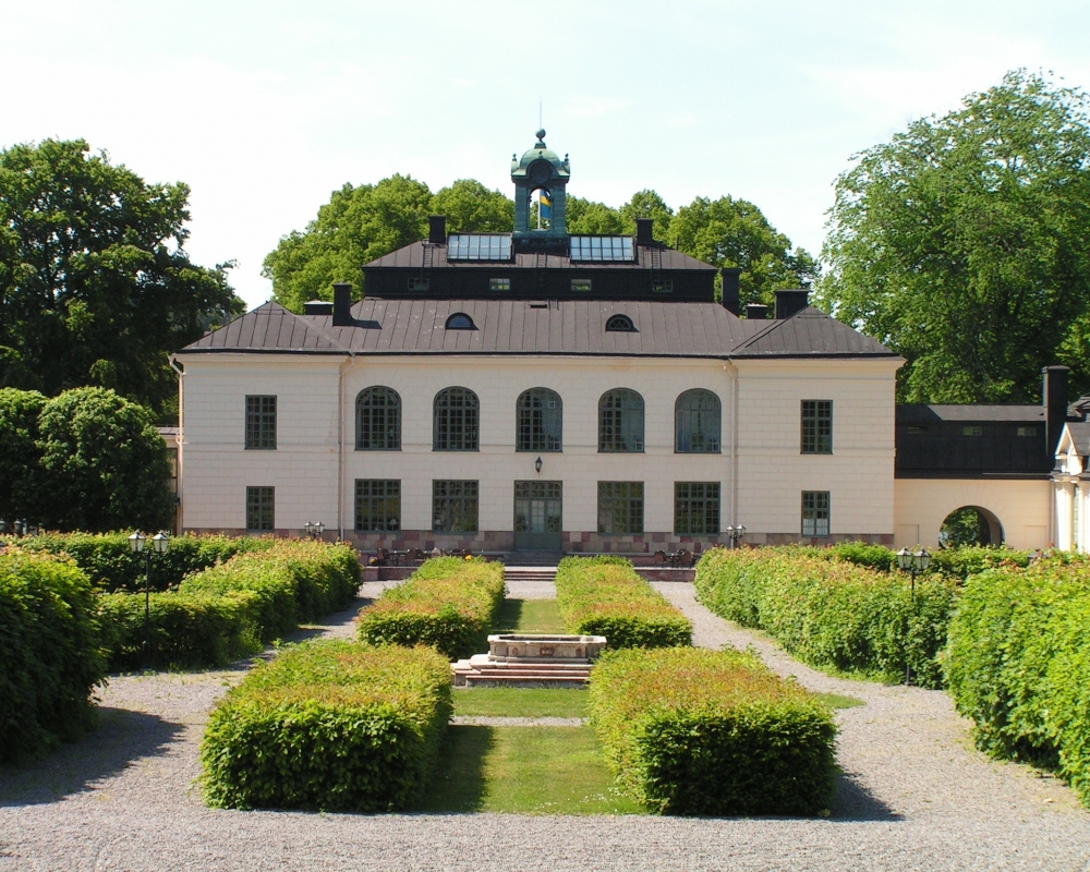 Le château de Näsby, un manoir blanc, vu à travers ses magnifiques jardins aux haies taillées et aux allées de gravier, sous un ciel d'été dégagé.
