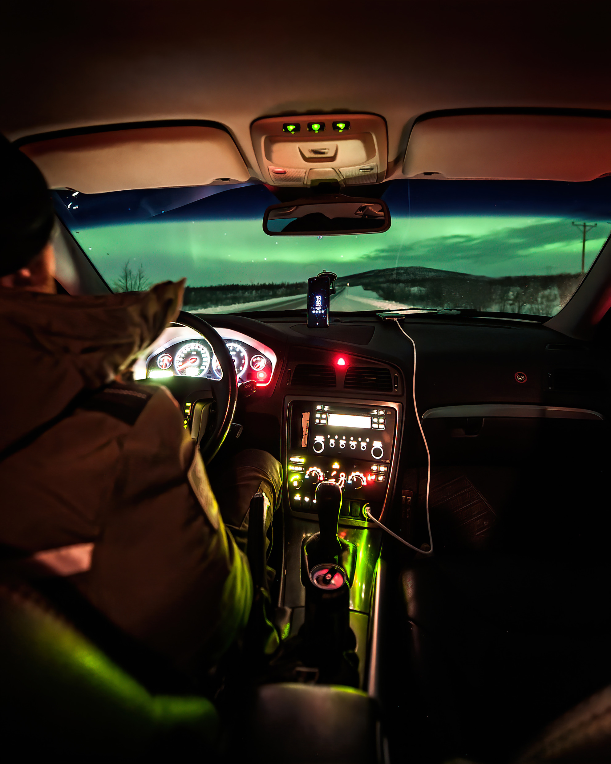 Een man die in een auto rijdt door een landschap met noorderlicht, gezien vanuit de auto.