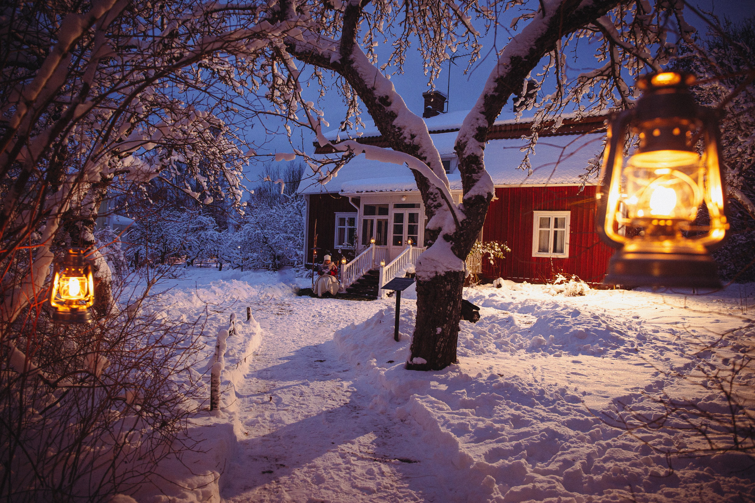A red cottage with white knots surrounded by snow. A woman is sitting on the steps of the house.