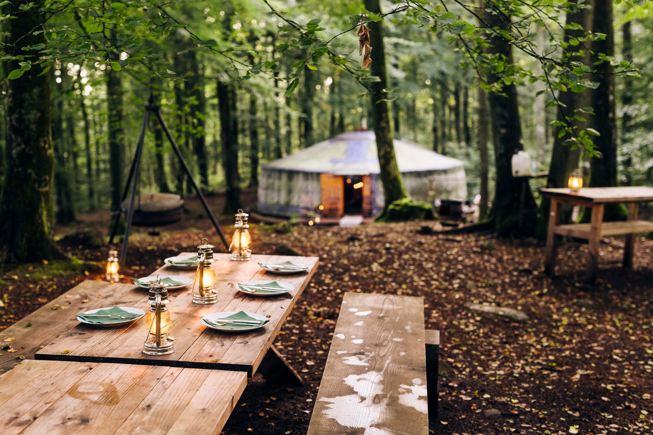 A wooden table with plates and lanterns stands in a forest. A large tent is in the background among the trees.