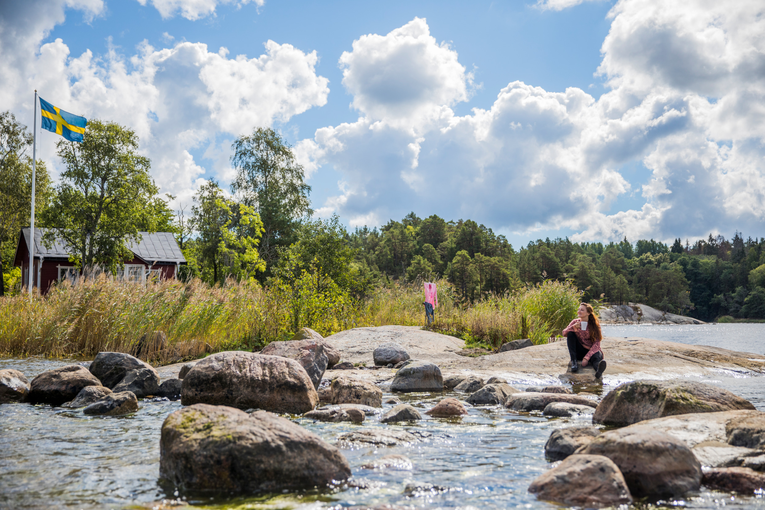 Eine Frau sitzt mit einer Tasse Kaffee auf einer Klippe am Wasser im Archipel. Ein rotes Häuschen und eine schwedische Flagge sind hinter ein paar Bäumen und Schilf versteckt.