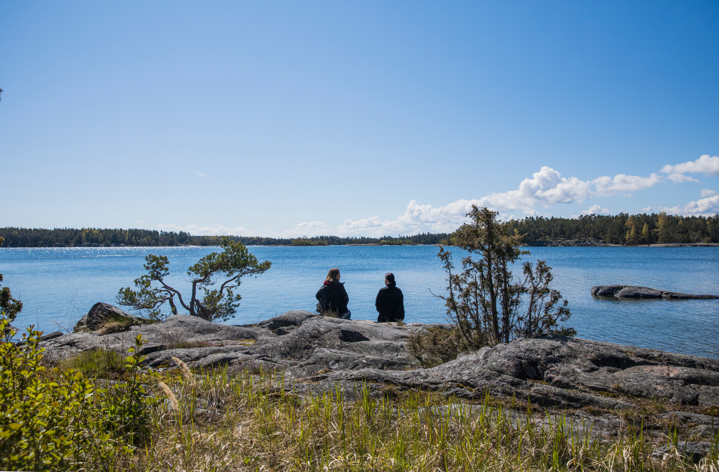 Two women sits on a cliff, with the back to the camera, enjoying the view of the sea in the archipelago. You see land further away.