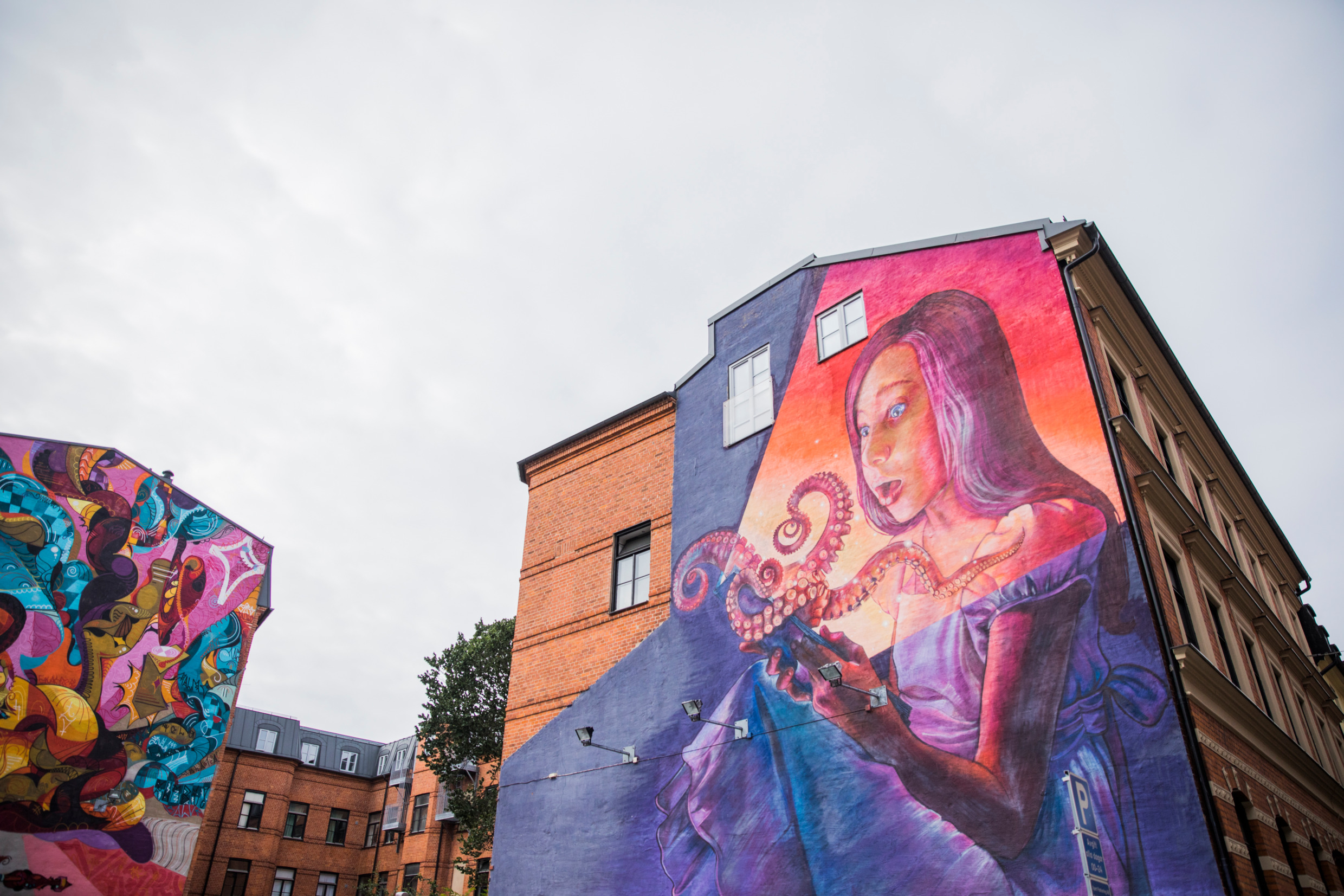 A mural painting on a house in Malmö depicting a woman holding an octopus and house beside you see parts of abstract painting.