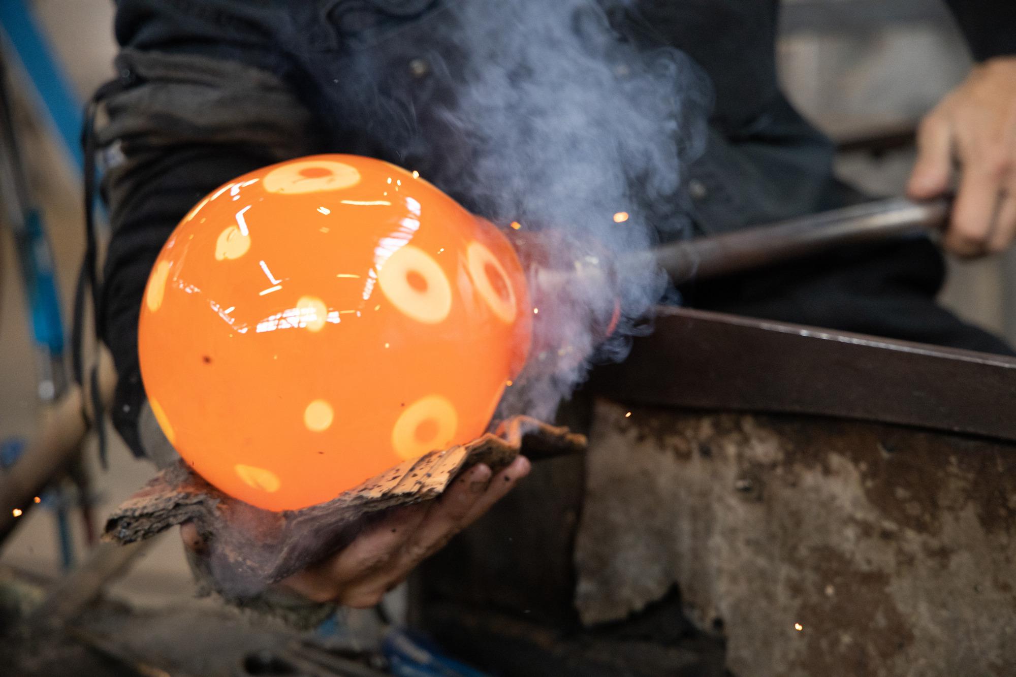 Gros plan sur une personne travaillant avec un verre soufflé en bouche. Le verre orange est chaud et brillant.