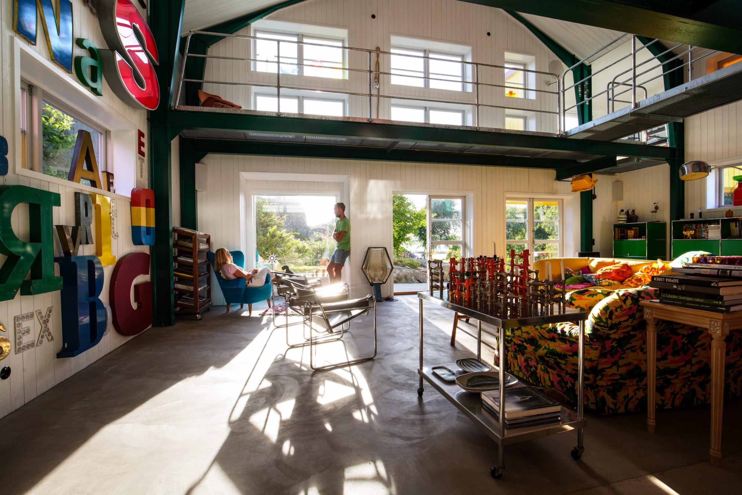 De zon schijnt in een woonkamer waar een persoon in een blauwe fauteuil zit en met een staande persoon praat.