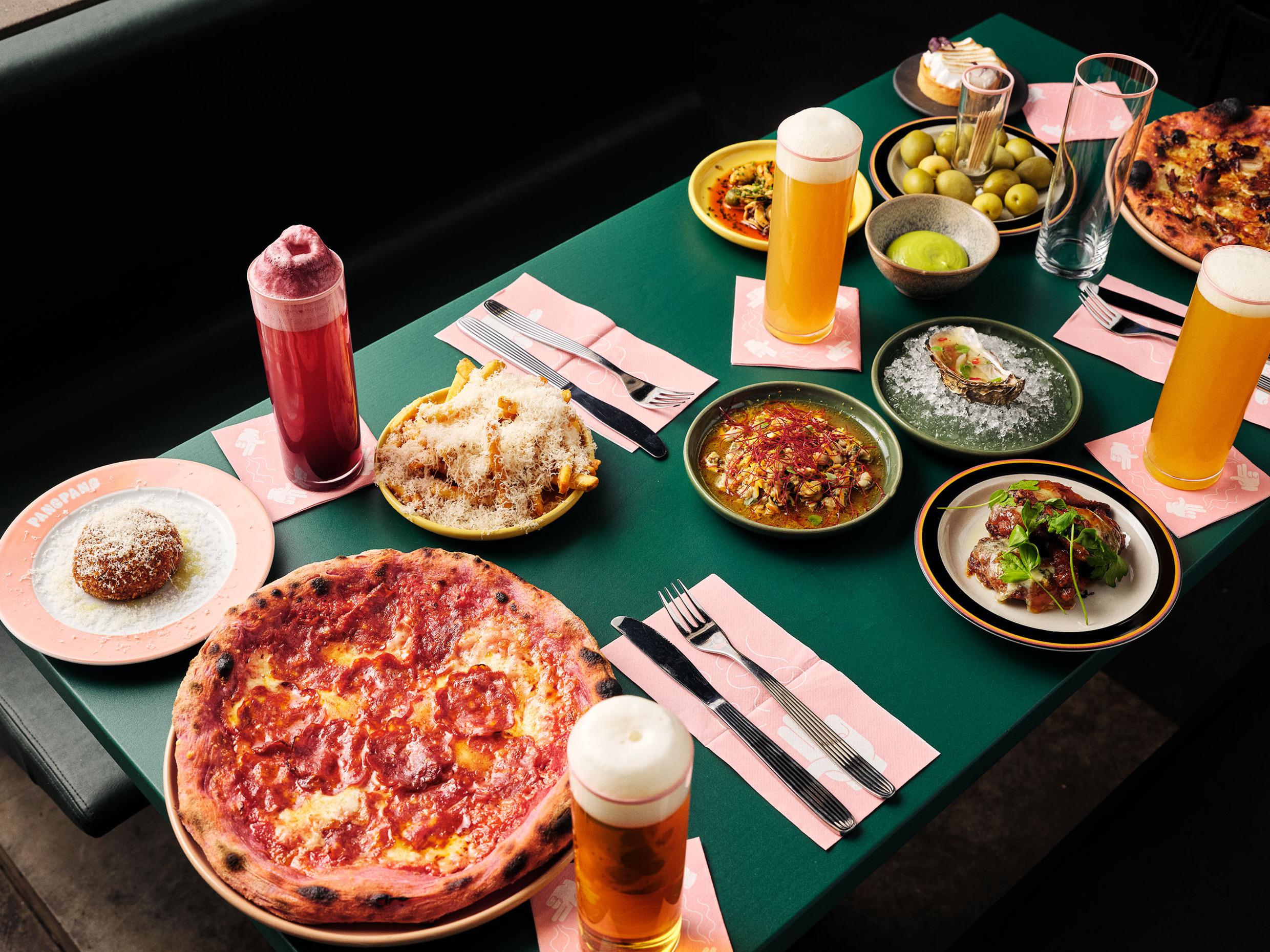 Een groene tafel met bestek, glazen gevuld met bier en borden met eten zoals pizza, frites, oesters en olijven.