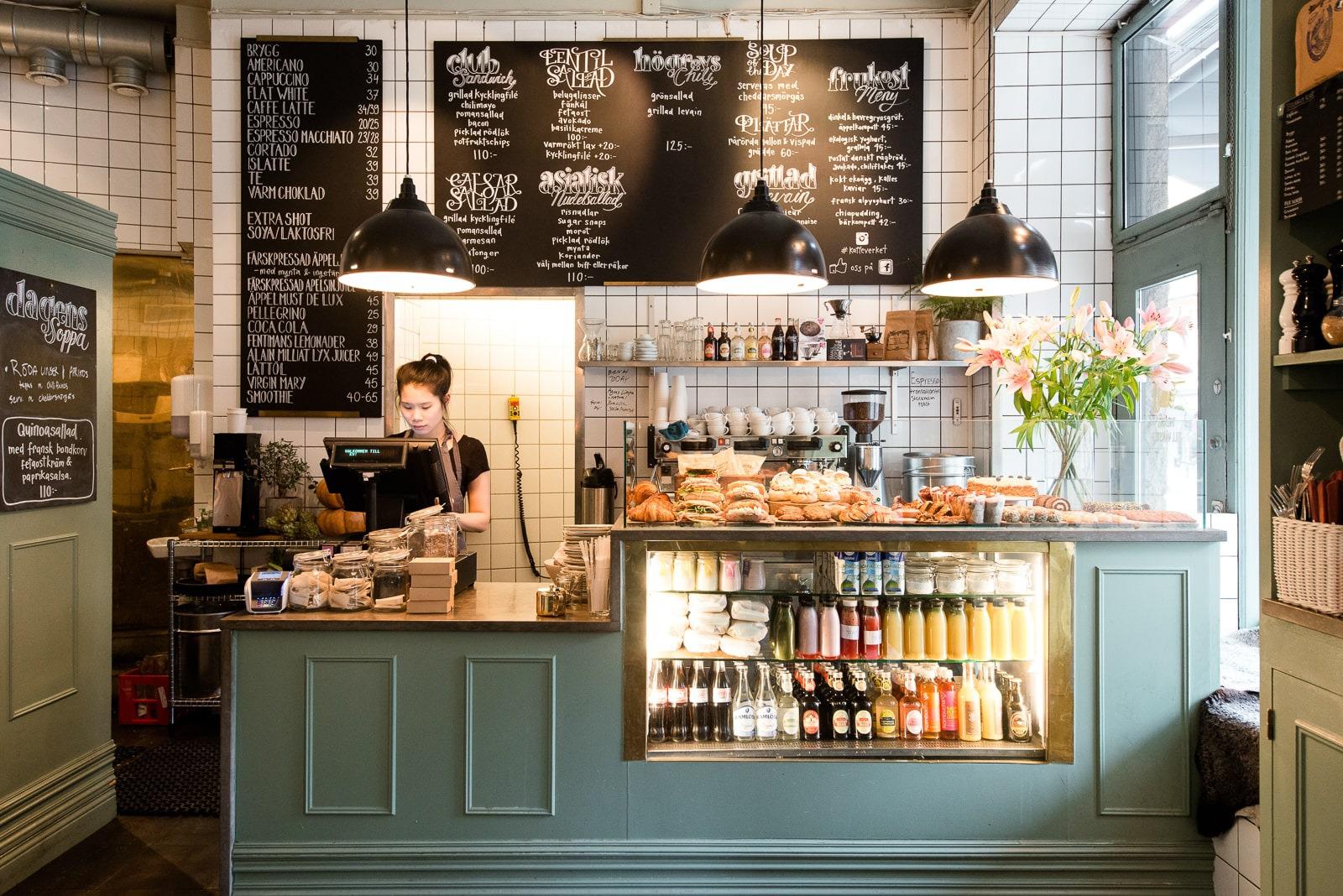 A waitress stands behind a counter in a café. There are blackboards with the menu written on them behind her and cookies and juices are on display on the counter.