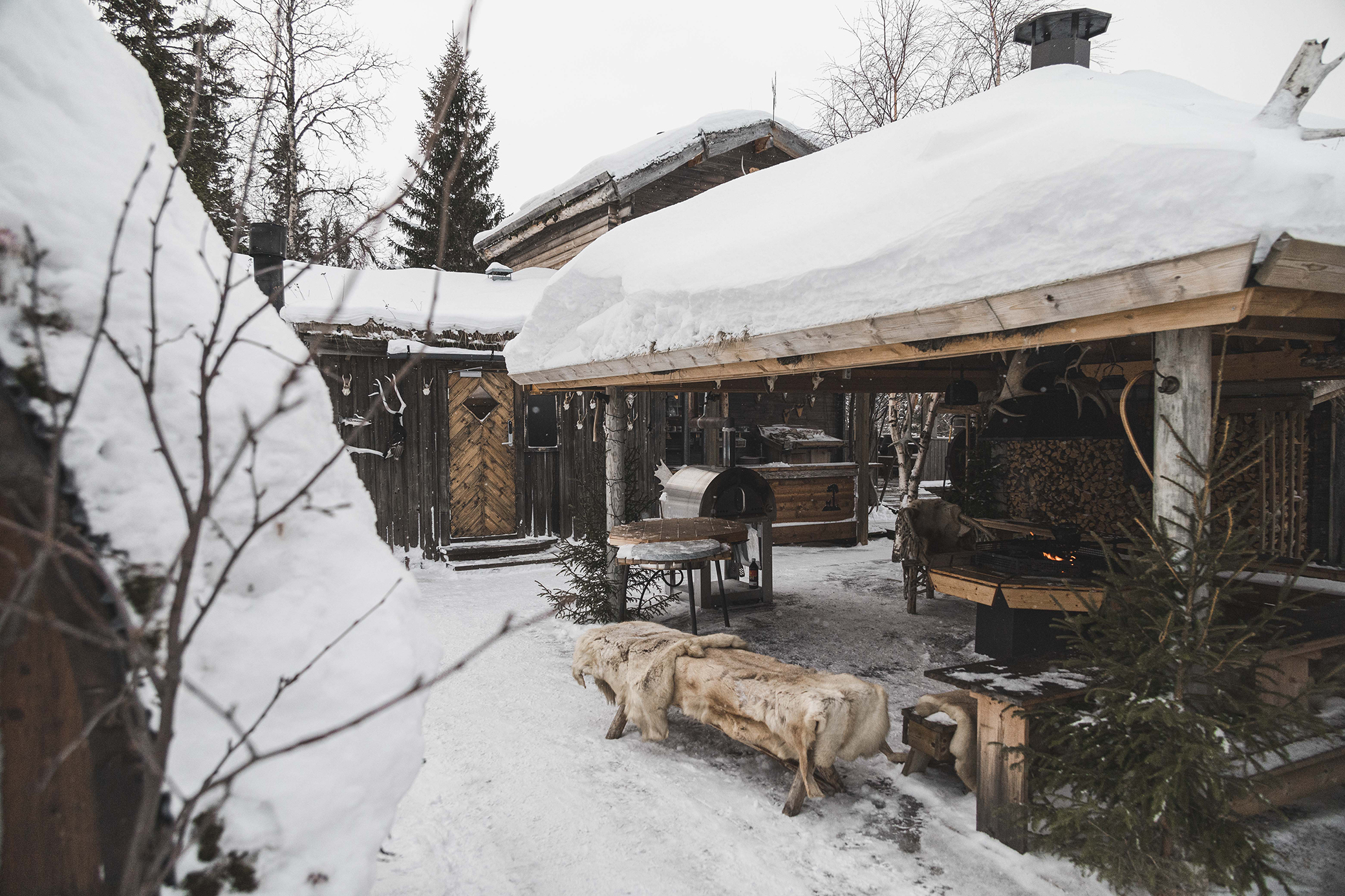 A rustic outdoor cooking area in winter with wooden buildings, a fire pit and snow-covered surroundings.