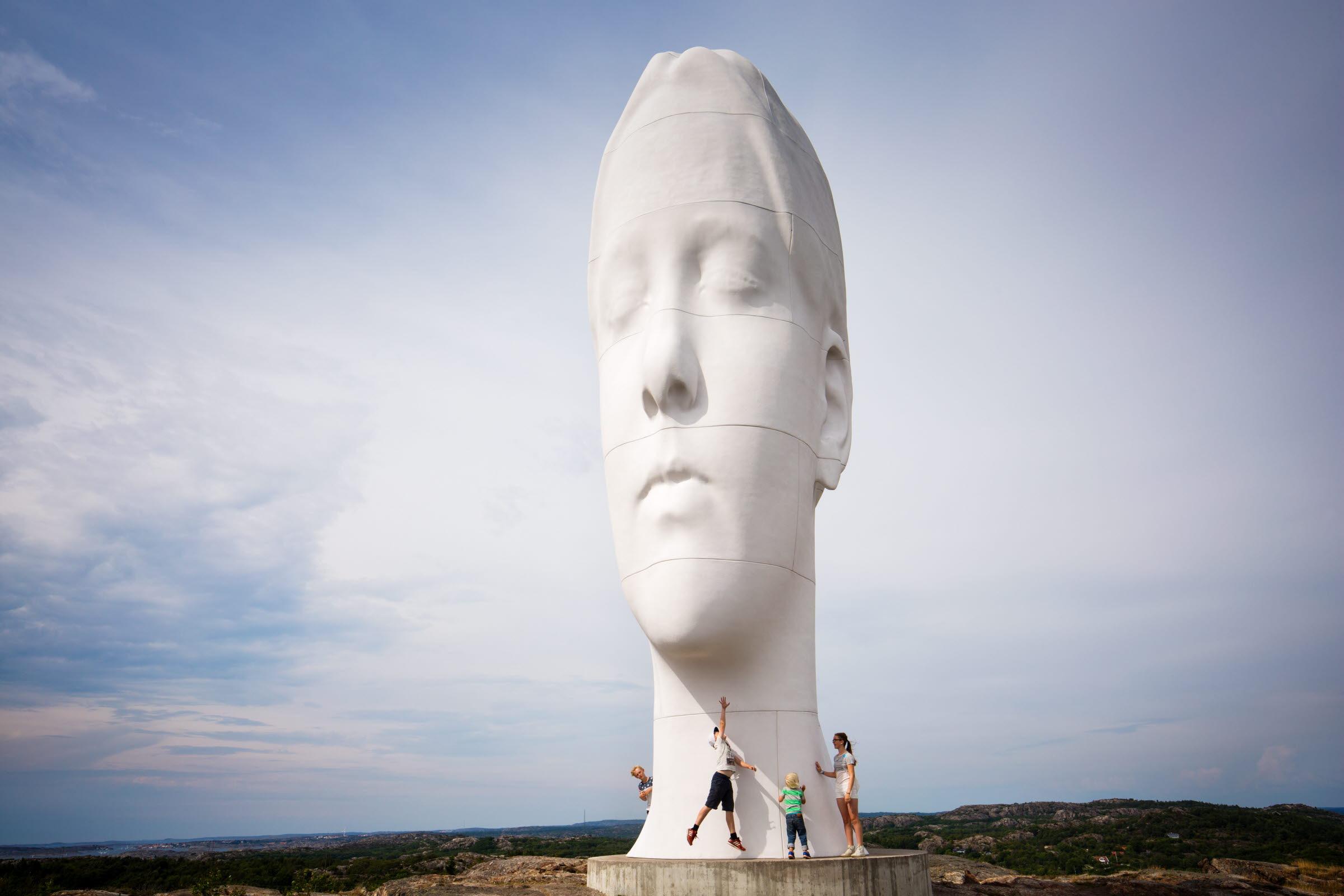 Eine große Statue in Form eines menschlichen Kopfes, die mitten in der Natur steht, und vier Menschen, die direkt darunter stehen.