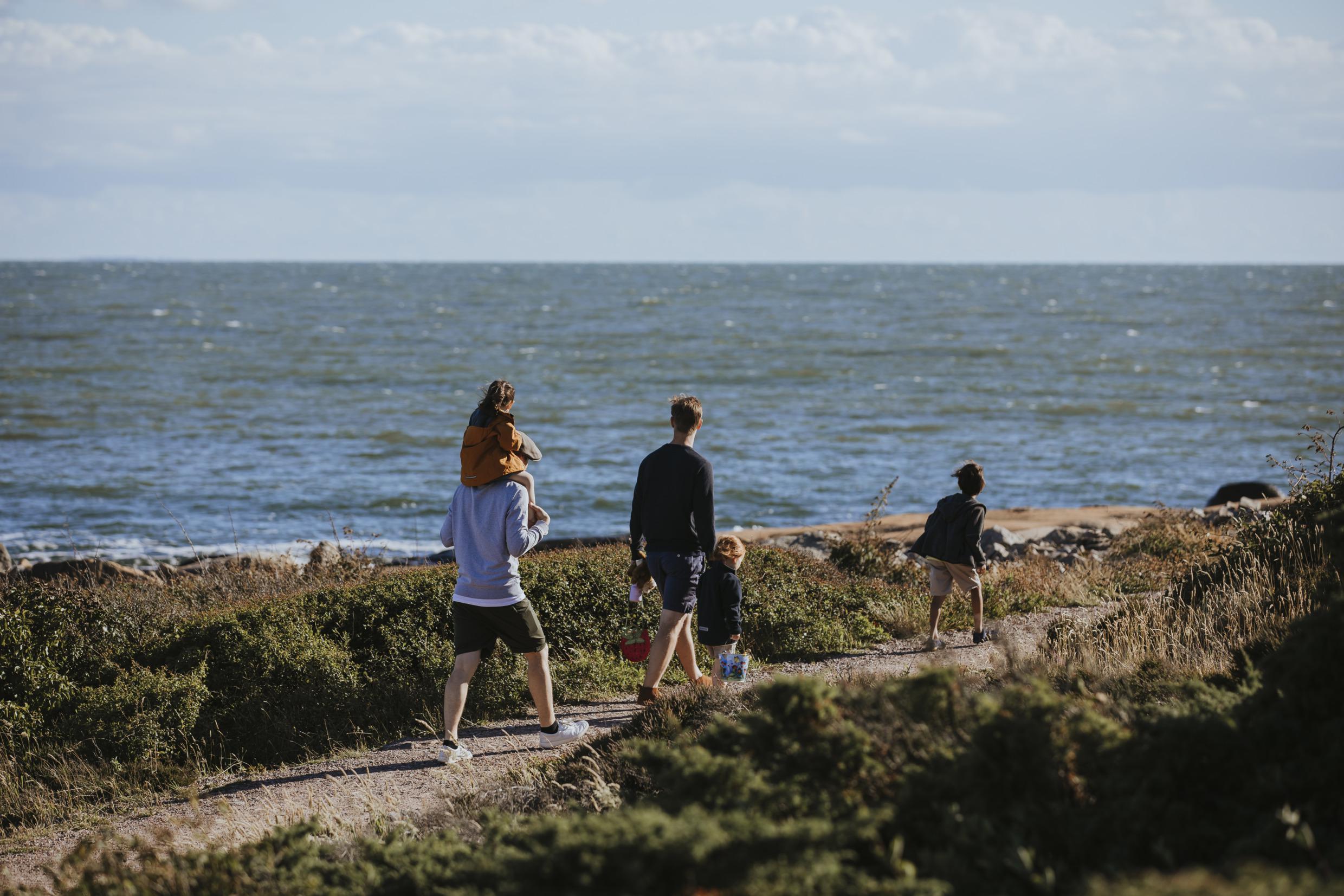 Twee mannen en drie kinderen lopen met de rug naar de camera, op een pad langs de kust. Een man draagt ​​een kind op zijn schouders.