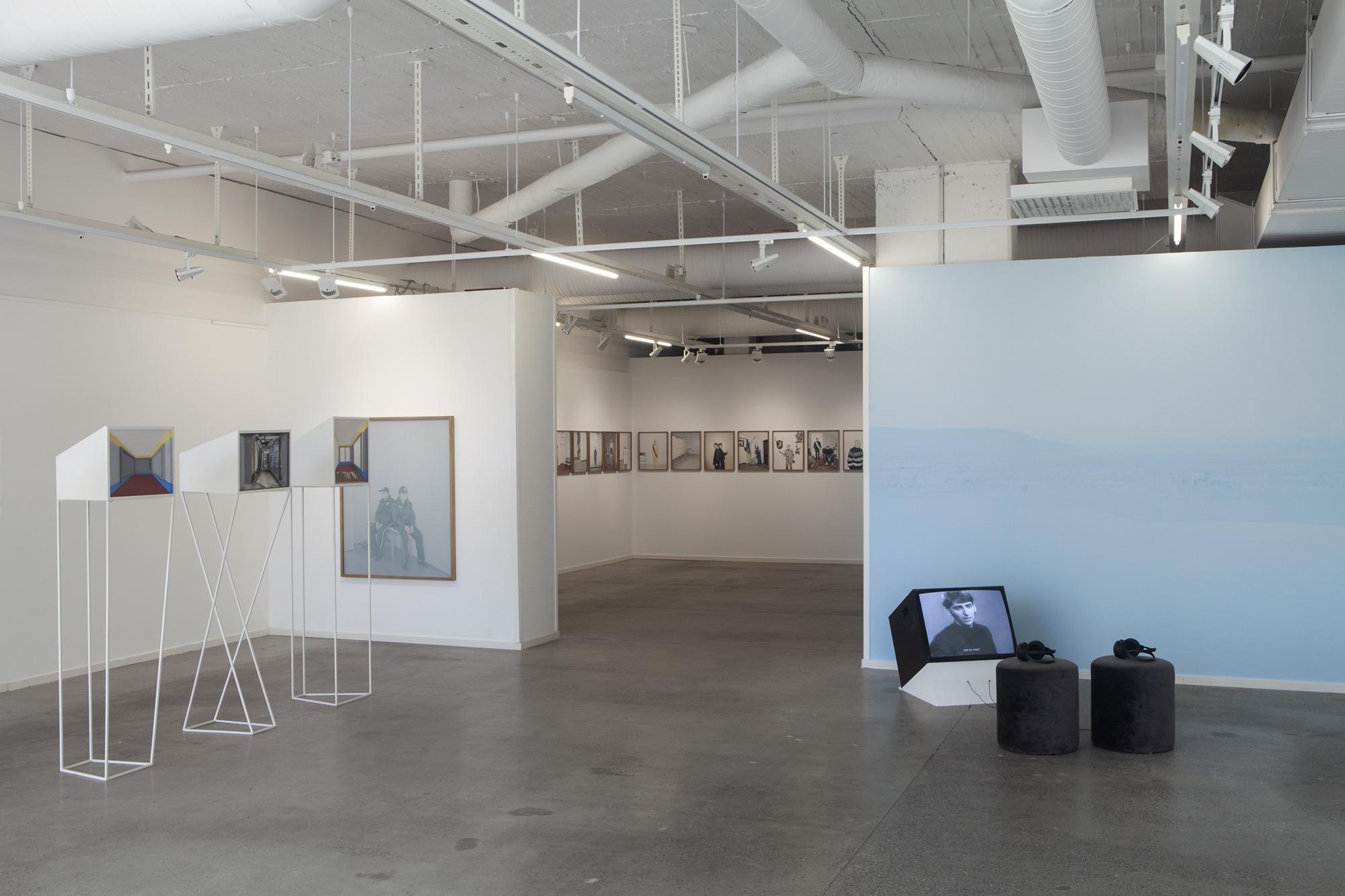 À l'intérieur d'une galerie d'art exposant des photographies d'art et des photographies documentaires. A droite se trouve un écran, deux poufs et des écouteurs.