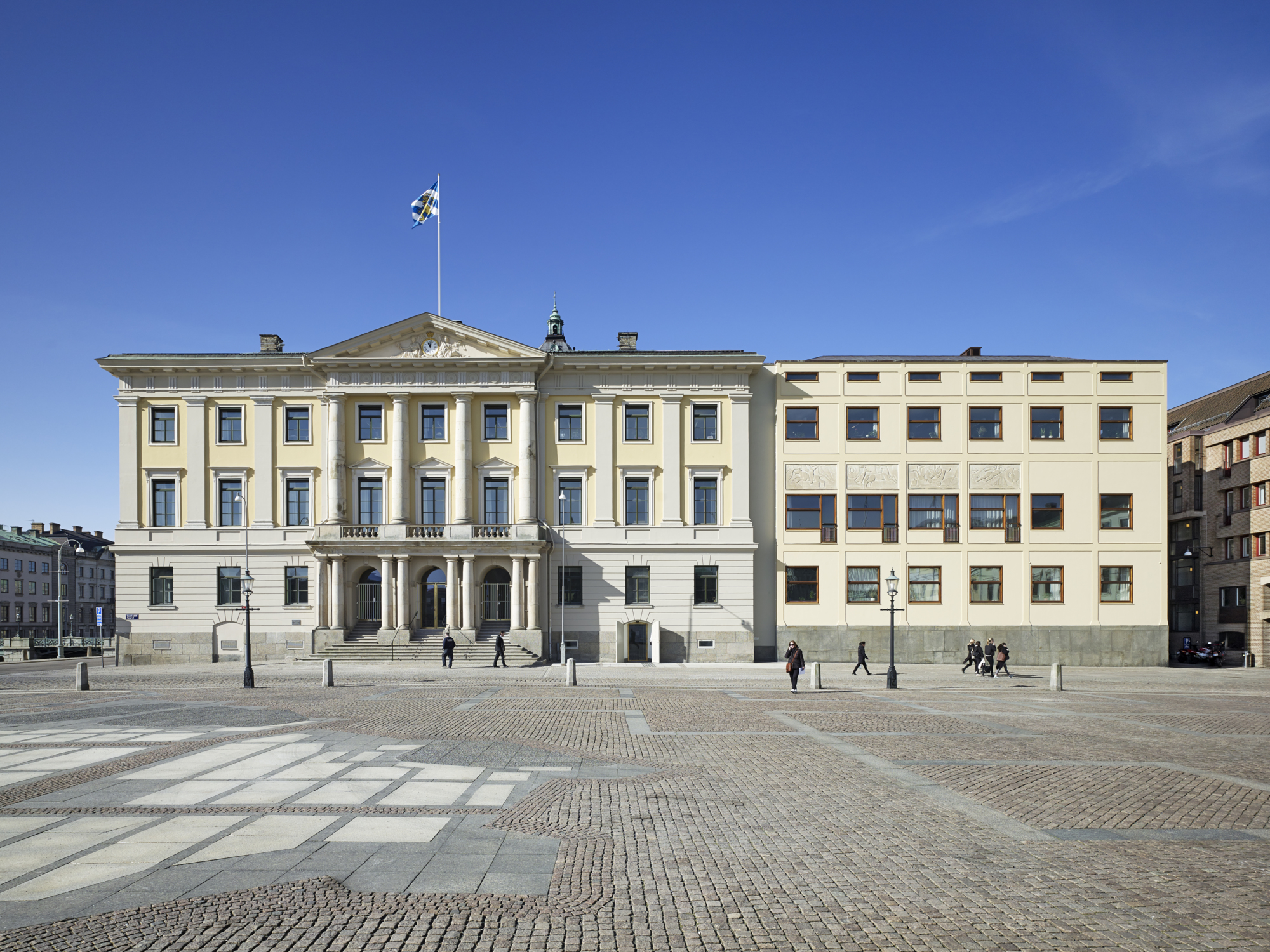 A large light yellow building with pillars, large sprouse windows and a big entrance, attached to a more modern building in the same colour.