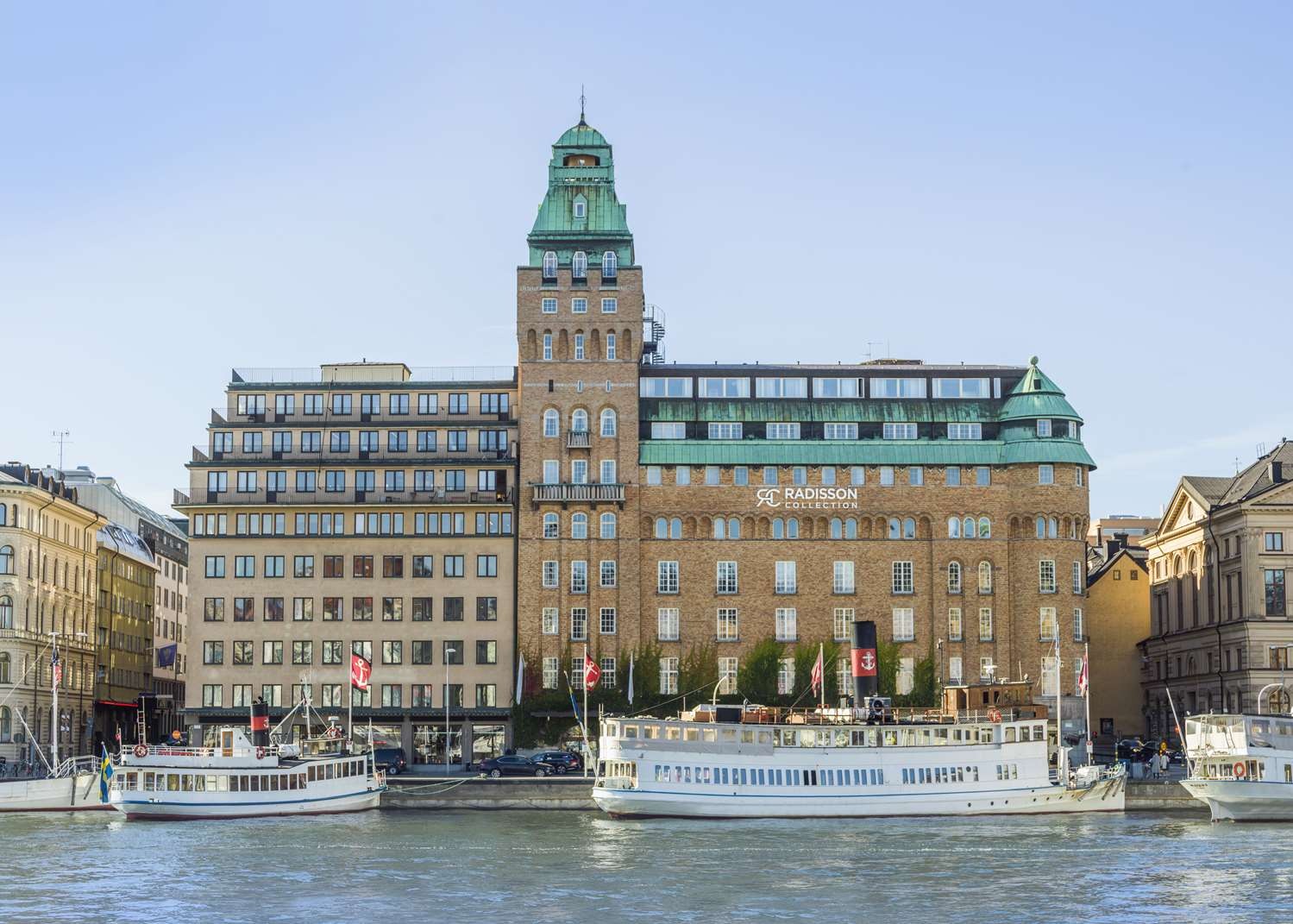Exterior view of Radisson Collection Strand Hotel in Stockholm, showcasing its brick facade, copper-roofed tower, and boats docked in the foreground at Nybroviken harbour.