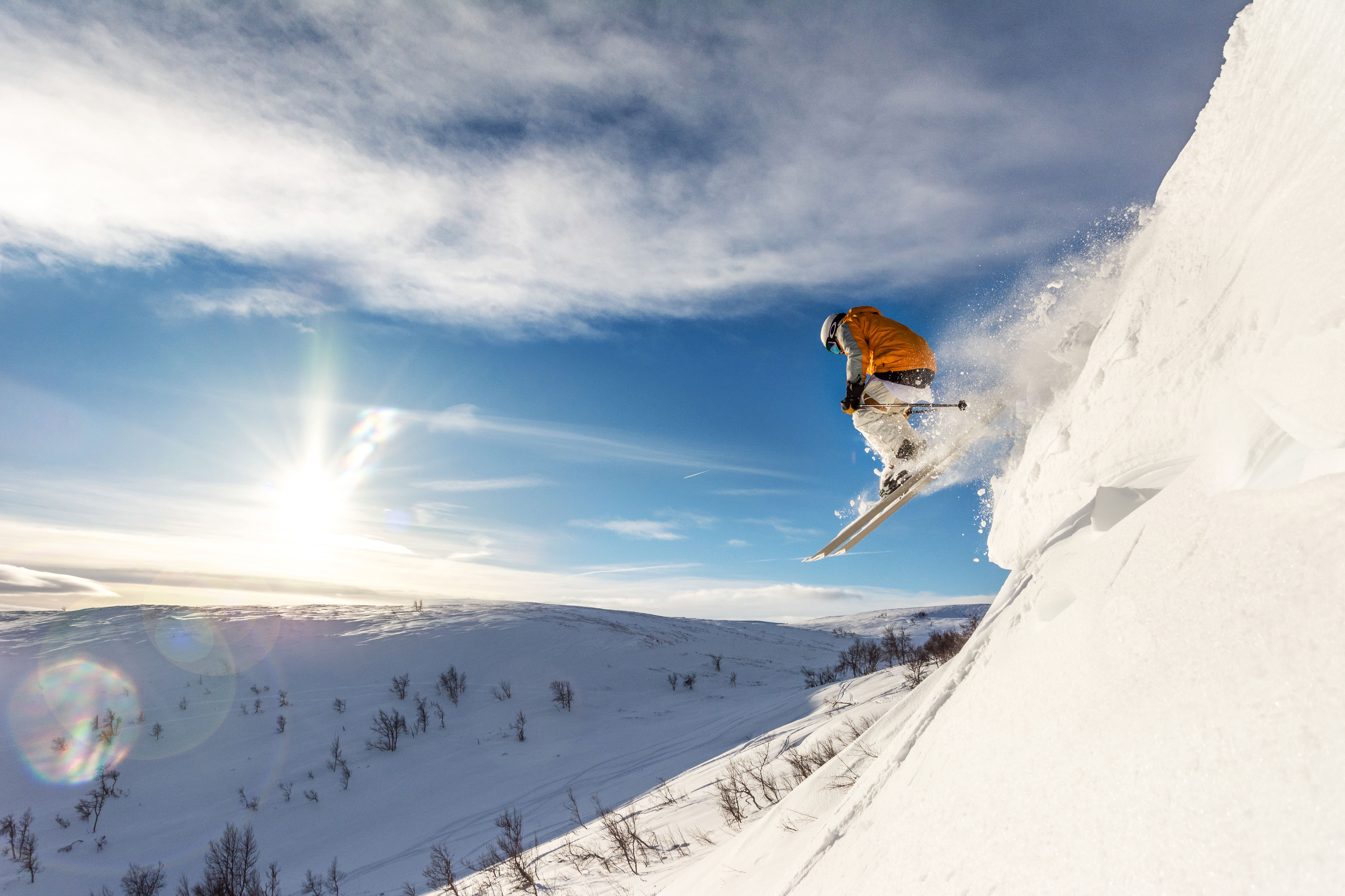 Skier drops into deep powder on a steep slope in Ramundberget, with sunlit mountains and scattered trees in the background.