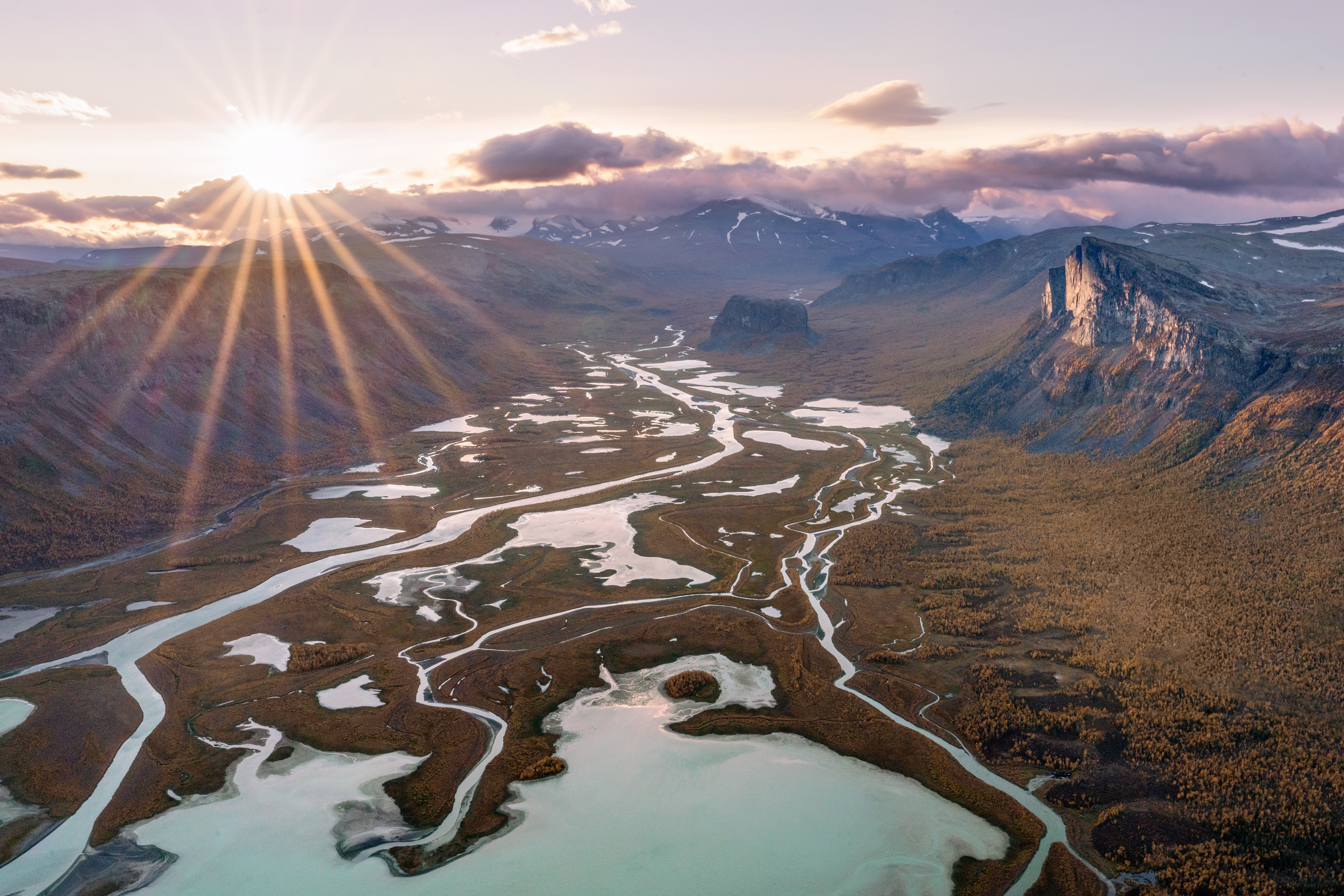 Sunlight breaks through the clouds over a vast mountain valley where braided rivers wind between wetlands and towering peaks.