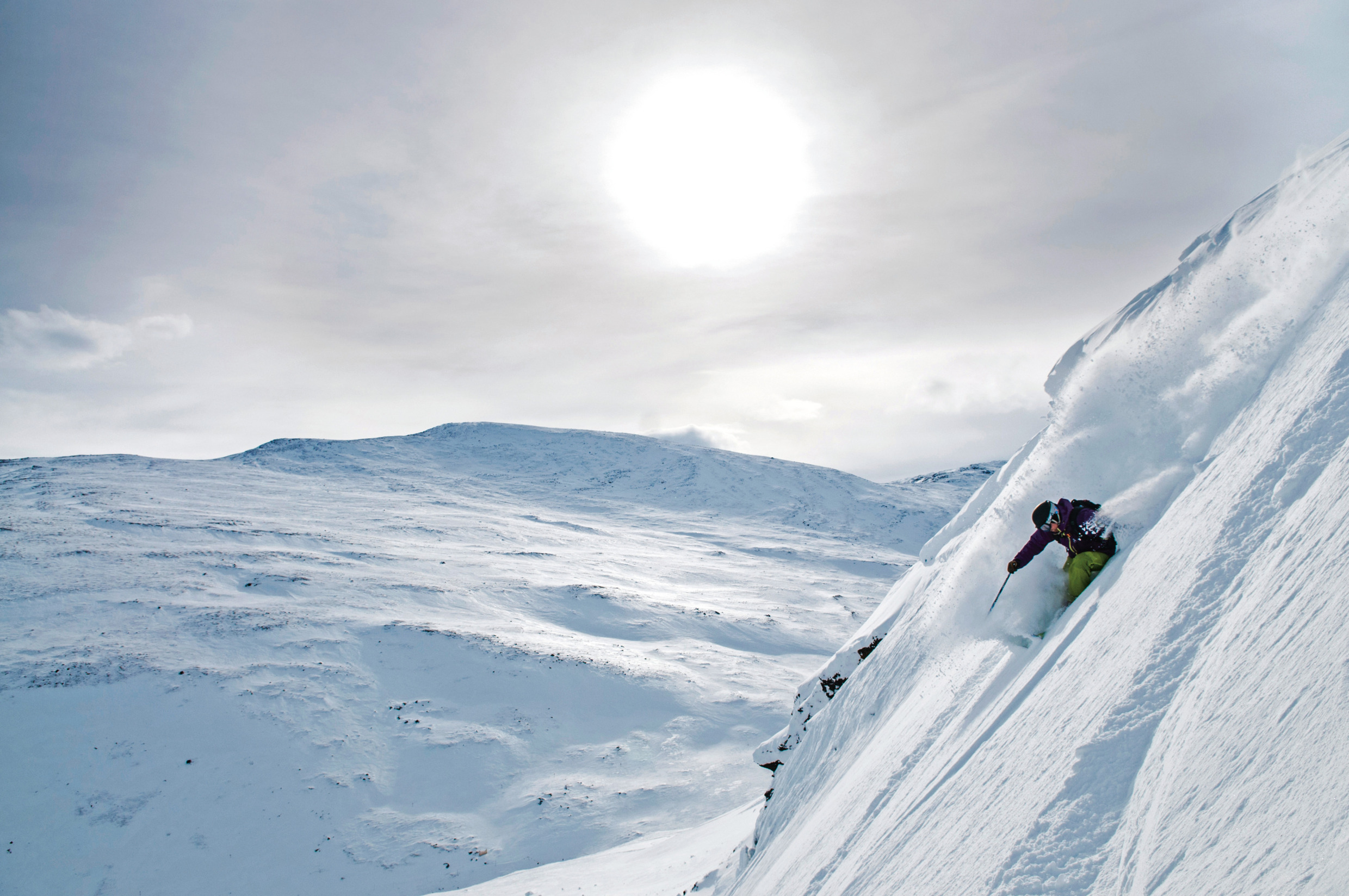A skier in bright gear descending a steep, snowy slope in Riksgränsen, with dramatic mountain scenery and a glowing sun overhead.