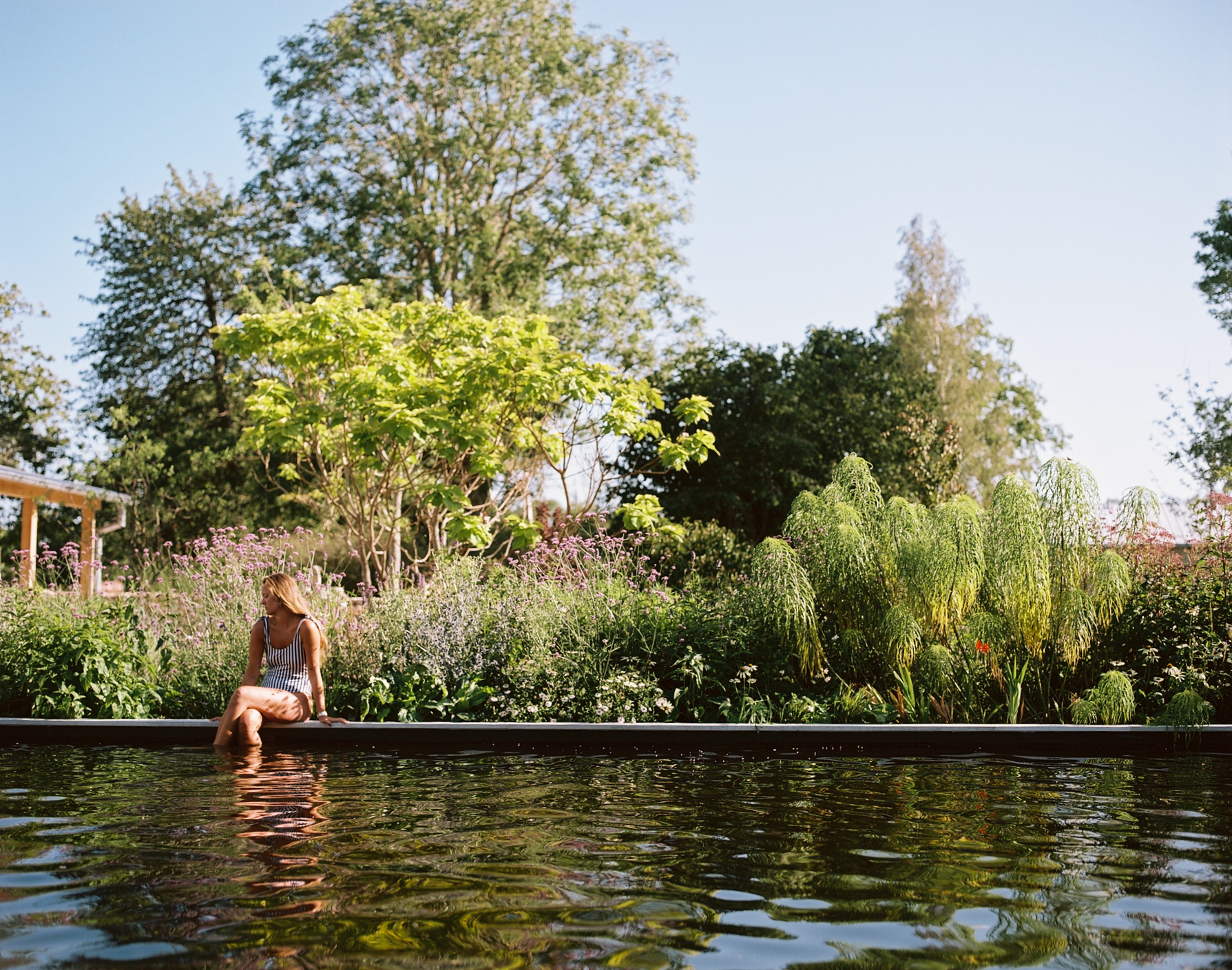 A woman sitting by a natural outdoor pool in a lush garden, with plants and trees in the background.