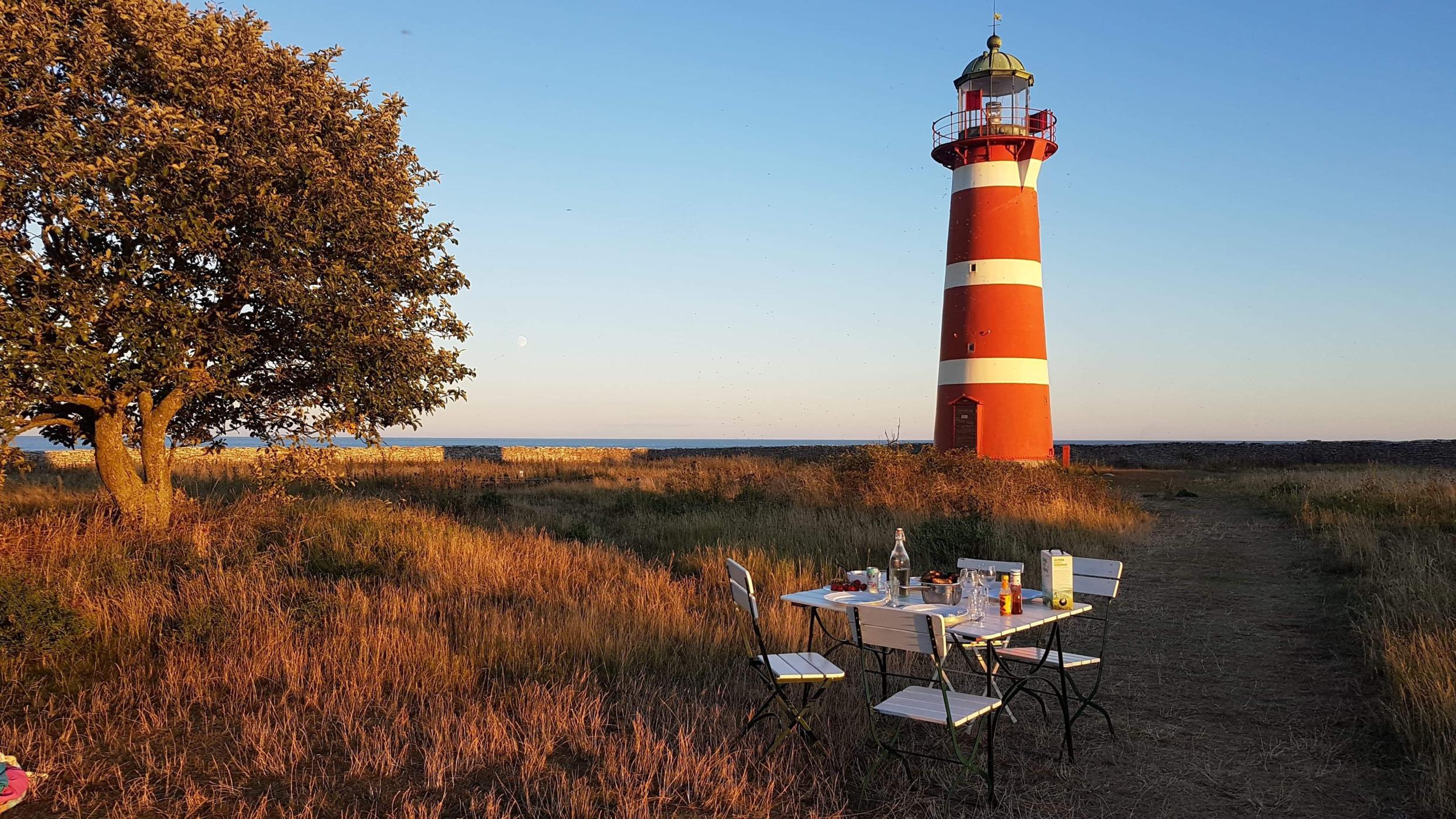 Ein Holztisch mit vier Stühlen steht im Sommer im Freien. Im Hintergrund ist ein rot-weiß gestreifter Leuchtturm zu sehen.