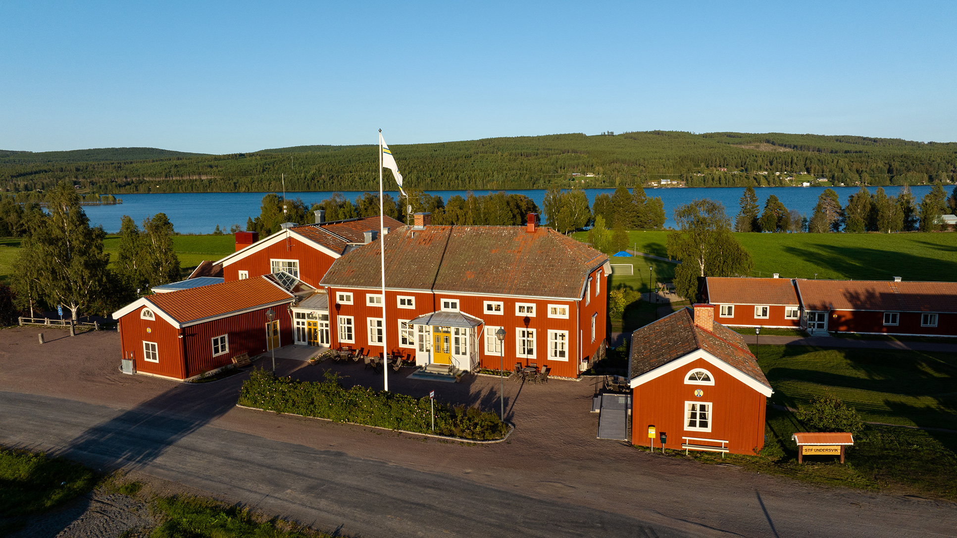 Red wooden buildings at STF Undersvik surrounded by fields, forest and a lake in the background.