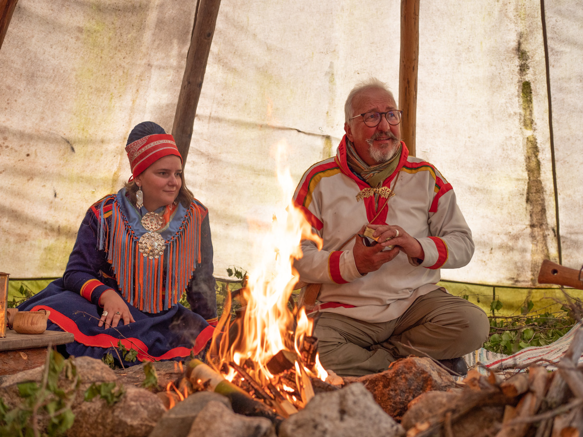 Zwei Sámi in traditioneller Kleidung sitzen an einem offenen Feuer in einem Lavvu, einem traditionellen Sámi-Zelt.